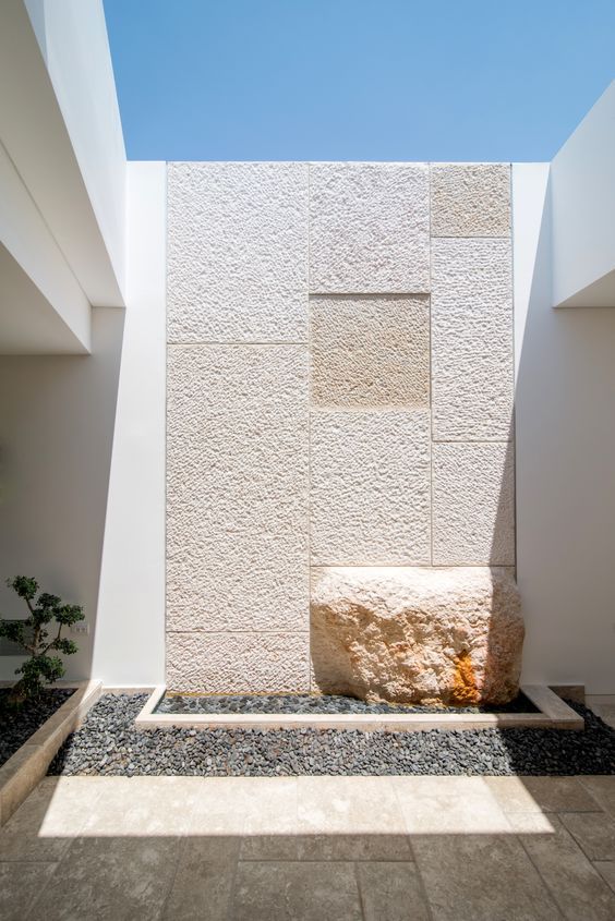 جدار داخلي مفتوح مصمم بتشكيلات هندسية من حجر الرياض الأبيض بنقشات متنوعة تتوسطه صخرة طبيعية بارزة كعنصر ديكور مودرن.