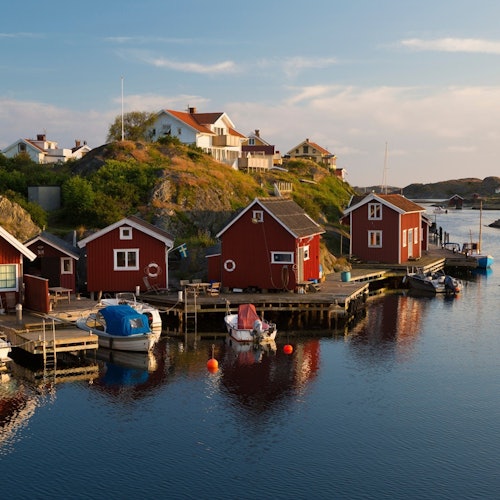 Red wooden houses and boats along a harbor with hills and more houses in the background under a clear sky.