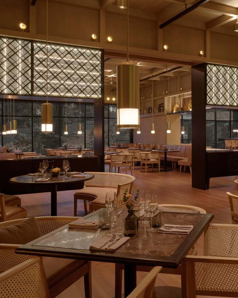 Interior view of a modern restaurant featuring wooden furniture, elegant lighting, and a cozy ambiance. Tables are set with glassware and cutlery, while large windows provide natural light.