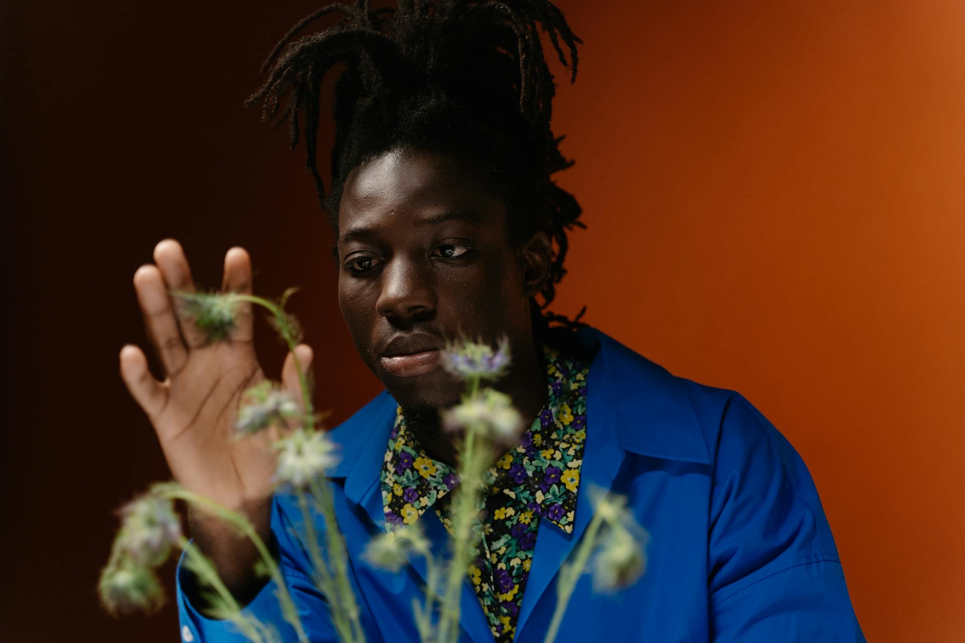 A man with dreadlocks wearing a bright blue jacket and a floral shirt looks thoughtfully at delicate green plants in front of an orange background.