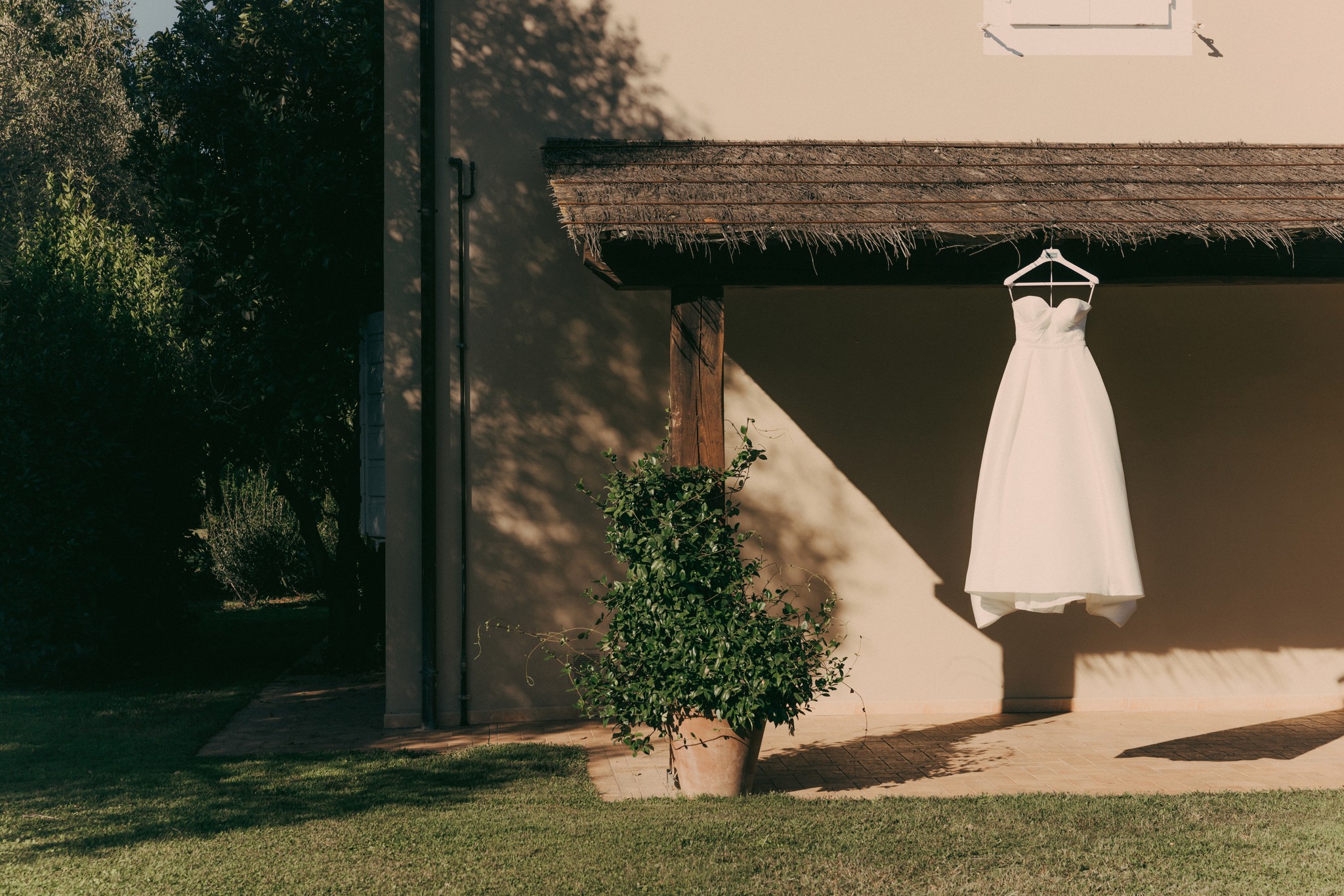 Abito da sposa appeso su muro rustico a Bolgheri, preparativi di matrimonio con luce naturale e ombre lunghe
