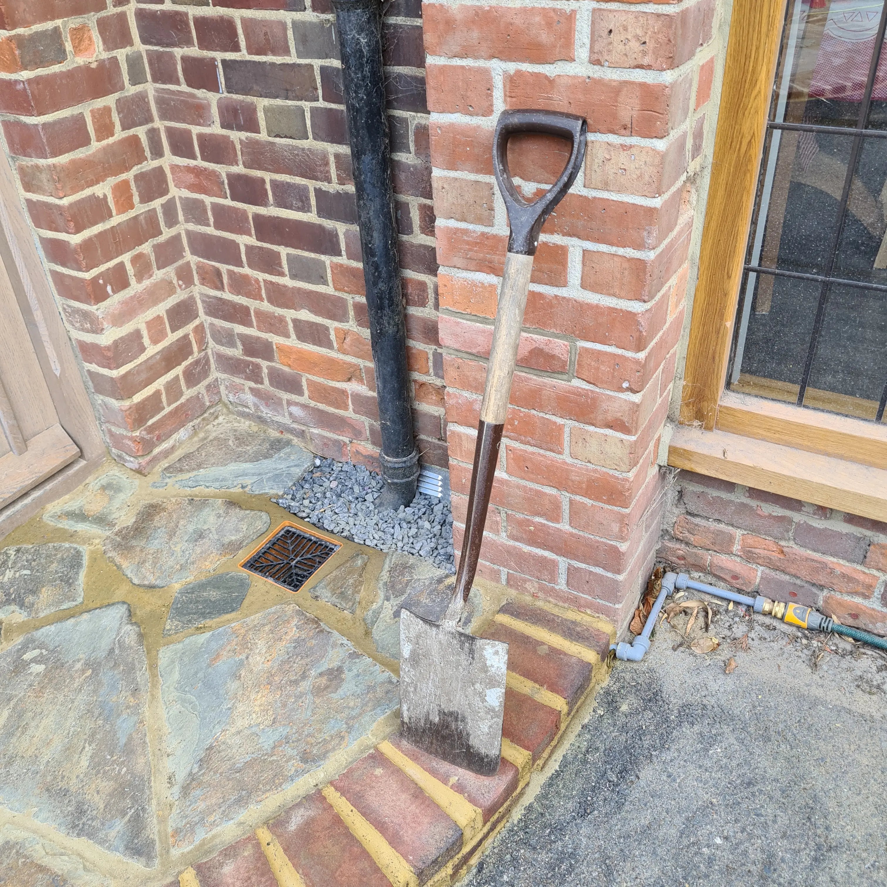 A brick wall corner with a shovel and a stone area at the base, next to a window.