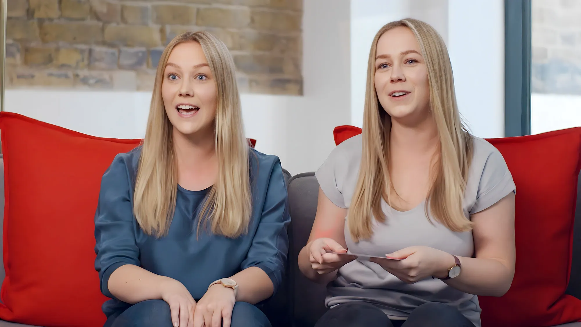 Blonde twins sitting on grey couch with red pillows