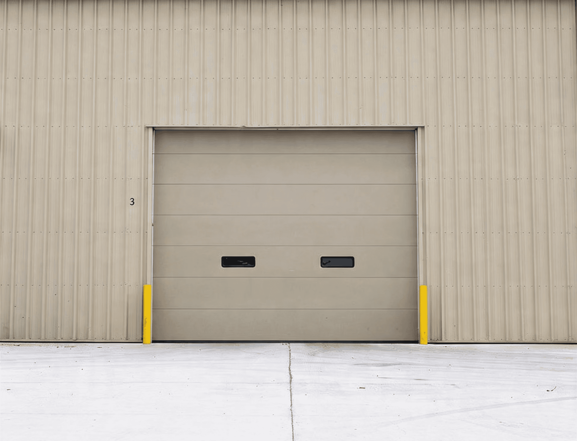 Tan industrial sectional garage door installed on a corrugated metal warehouse exterior. This heavy-duty commercial overhead door features two small vision windows and yellow safety bollards, optimized for high-traffic logistics and shipping facilities.
