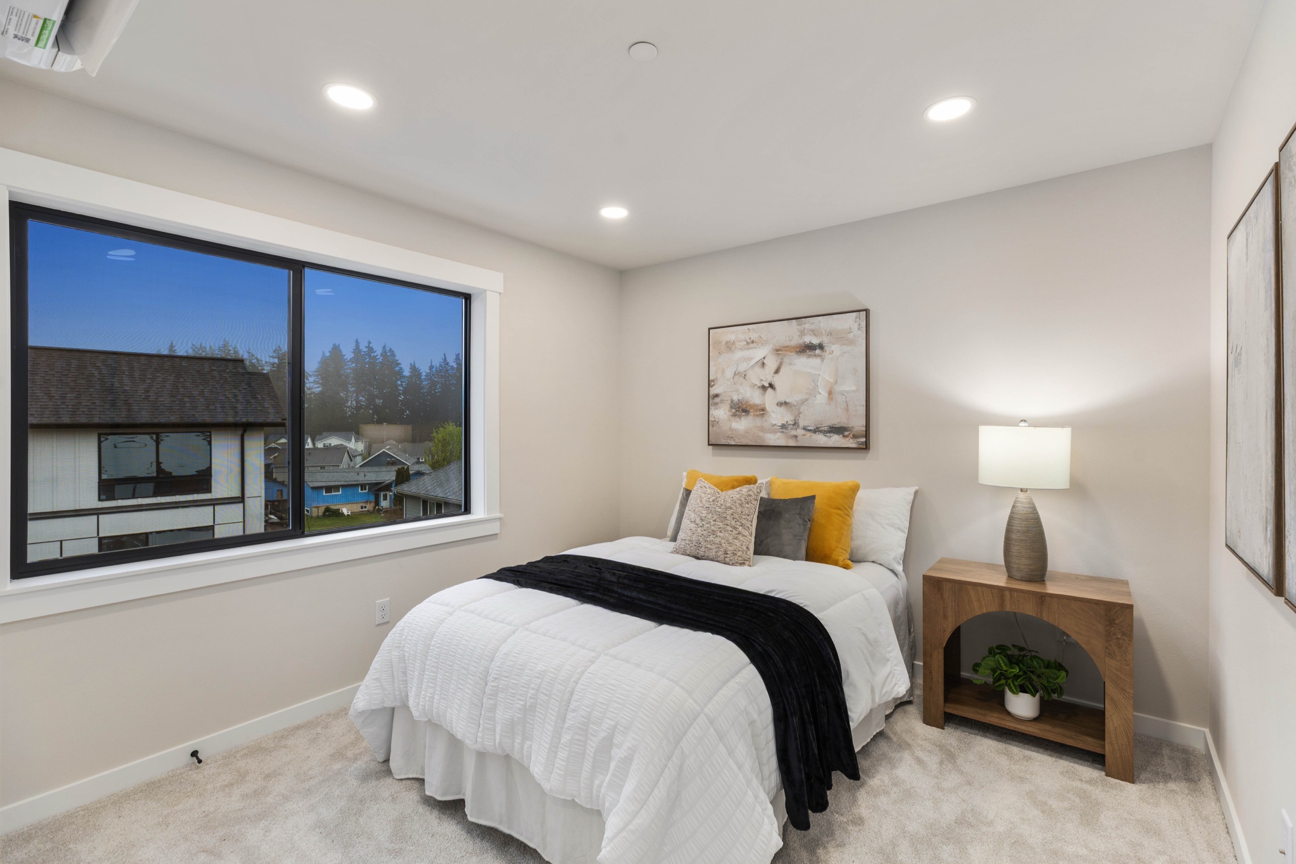 Airy luxury secondary bedroom with large window overlooking tree-lined street, plush carpet, wooden nightstand, table lamp, and neutral wall art