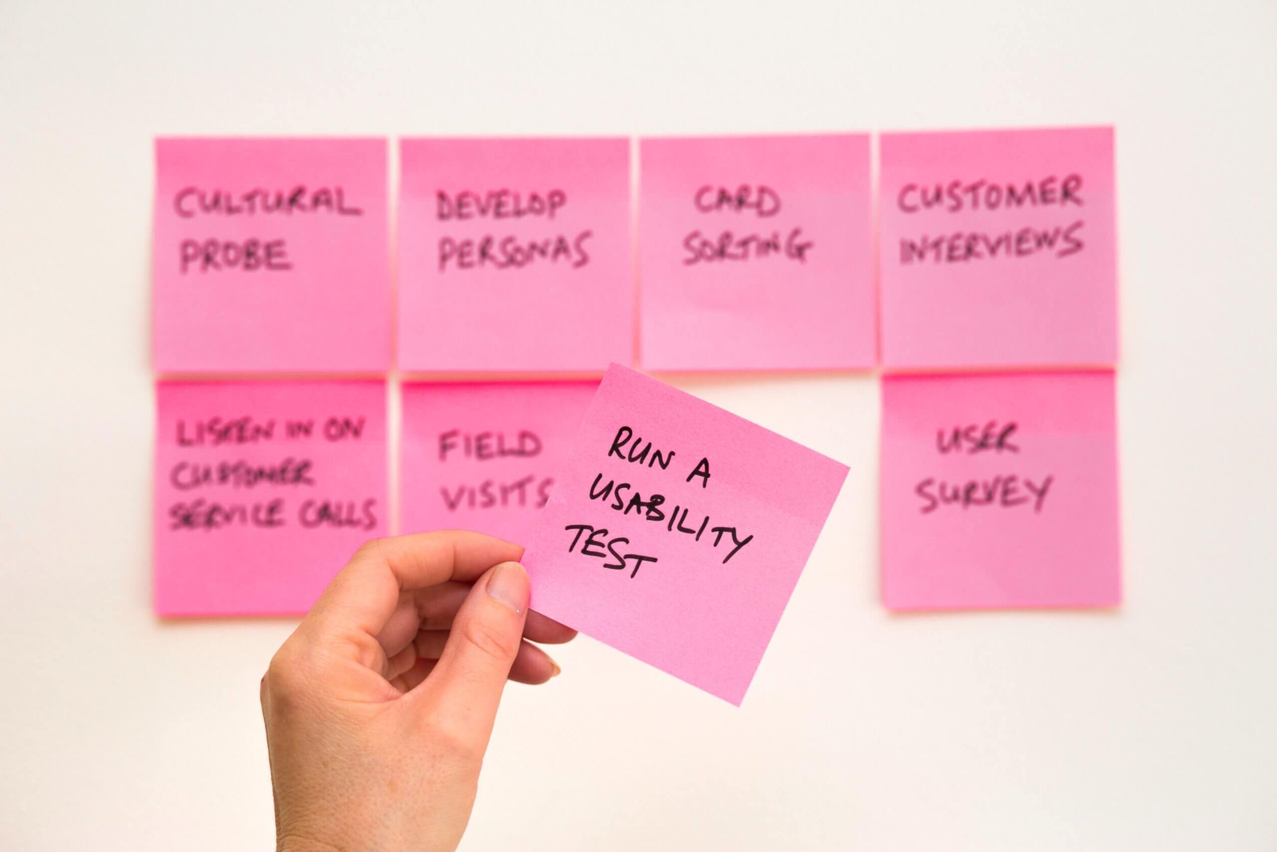 A hand holding up a sticky note that says "run a usability test", with other notes attached to a wall in the background