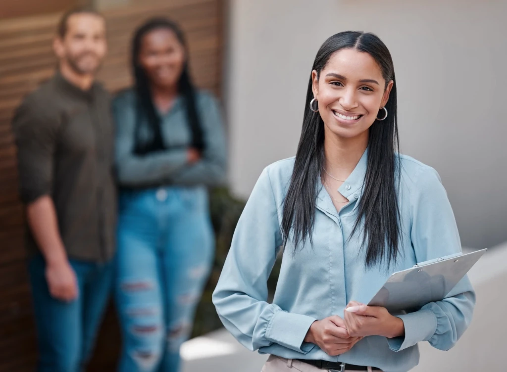 Smiling mixed race real estate agent holding a clipboard, with her clients behind her out of focus