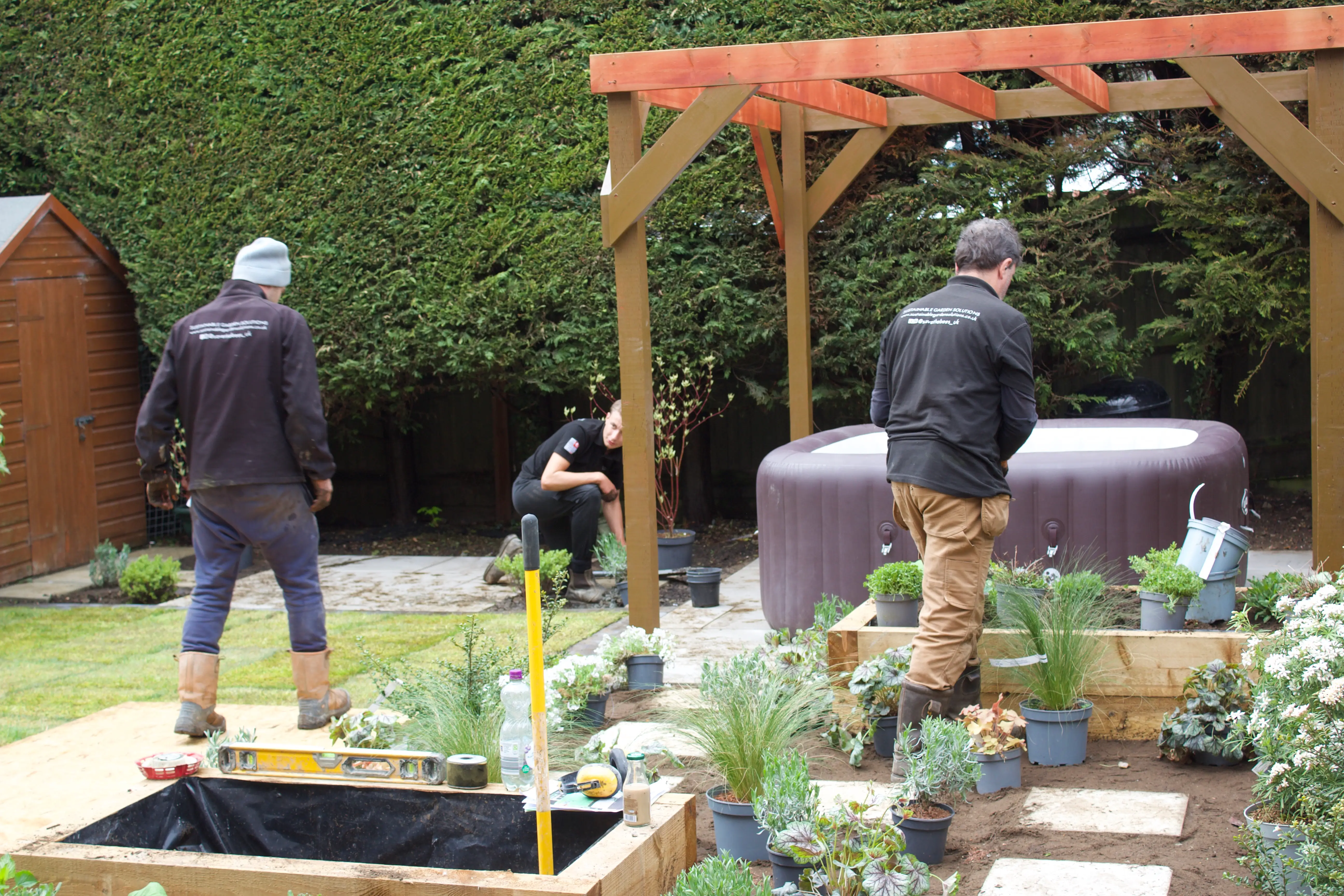 Two people in jackets stand in a garden with plants, near a wooden structure and a gray bench.