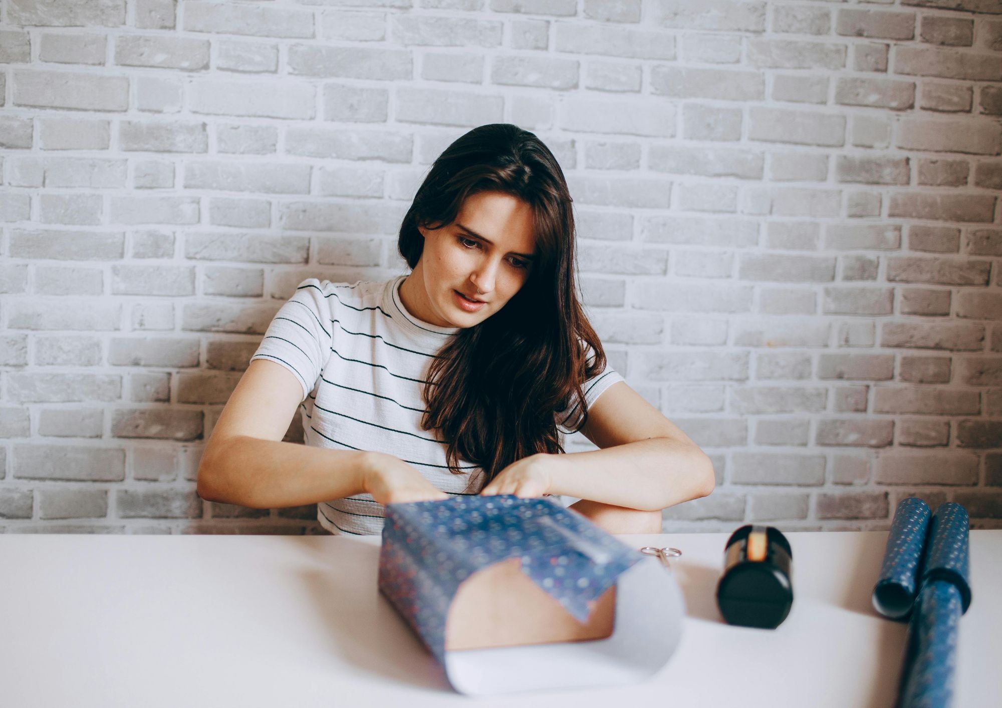 Mulher sentada à mesa embrulha uma caixa de presente com papel azul estampado.