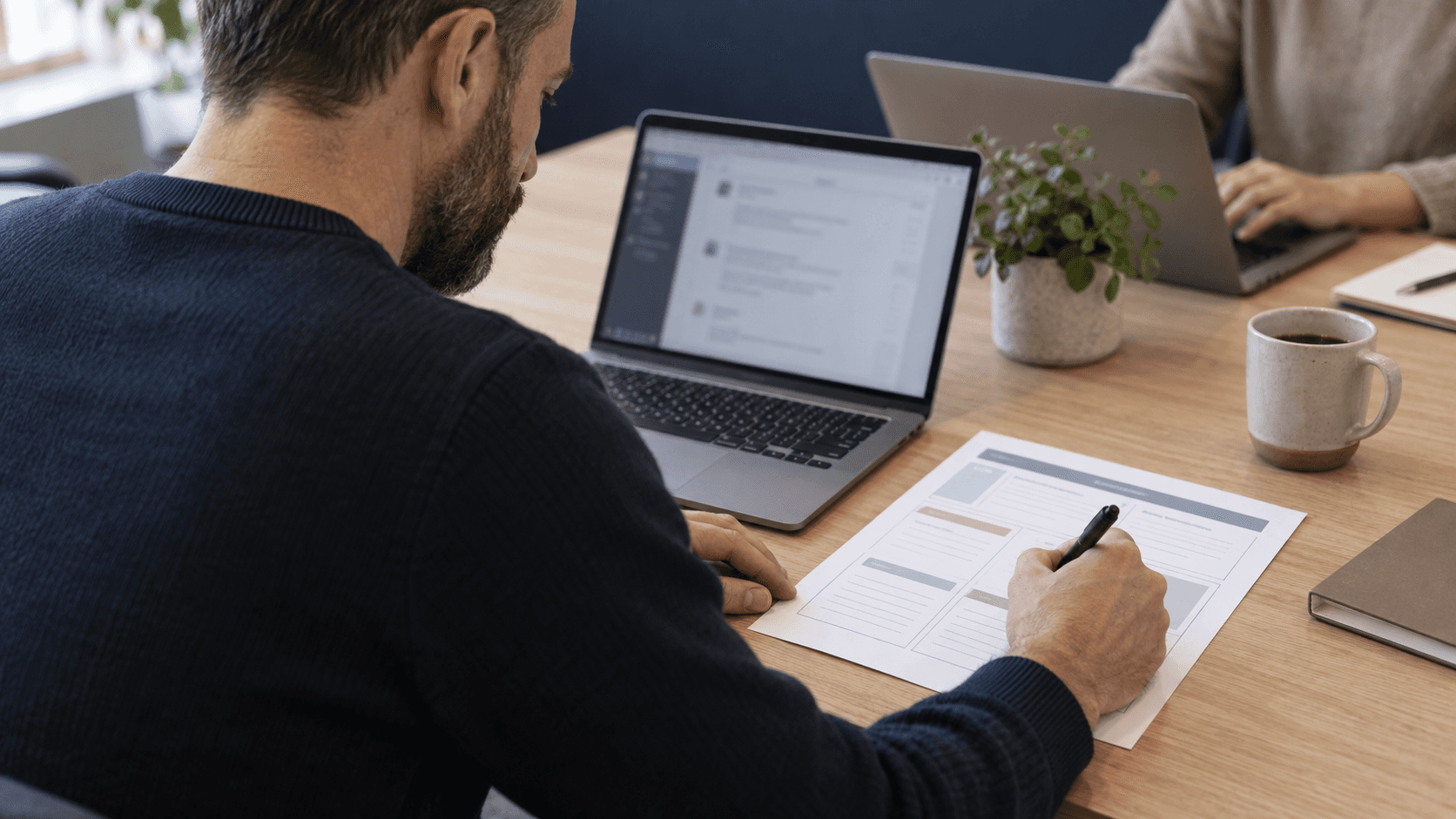 A professional fills in a printed prompt template beside his laptop while a colleague works across the table