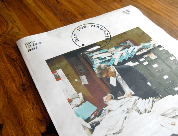 A folded magazine lies on a dark wooden surface. The cover displays a circular logo reading “DAY JOB MAGAZINE” with a dot in the center. The cover also features the text “Issue No Zero free!” and “Everyday Working Life” in the top right corner. A man in a suit is pictured on the cover, leaning over a desk with papers and files.
