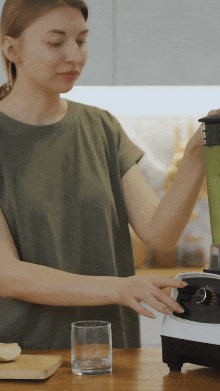 Woman influencer in a kitchen blending a green smoothie in a blender while standing near a wooden counter with a glass and a cut apple. She wears a green t-shirt and operates the blender with a focused expression, demonstrating a healthy lifestyle routine.