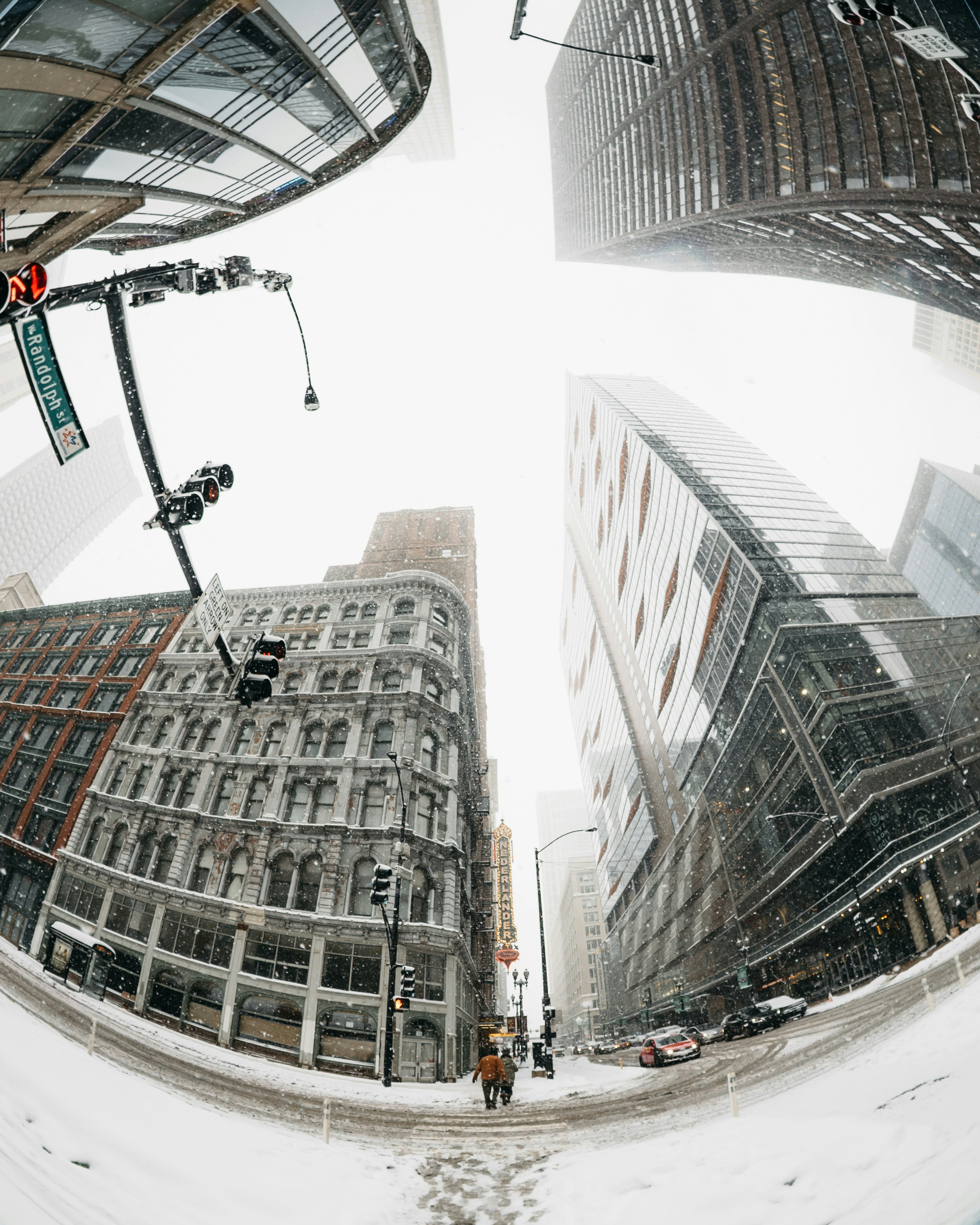 Snow falls on a city street with tall buildings.