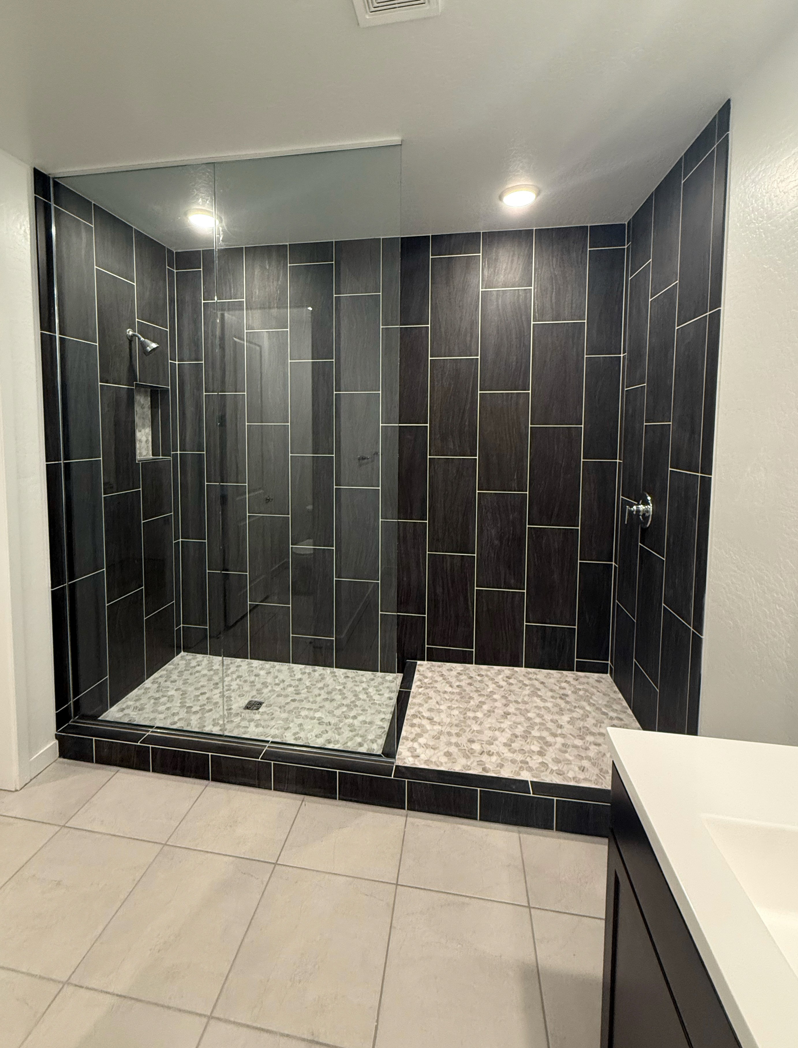 Spacious walk-in shower with dark grey rectangular wall tiles, a contrasting light pebble mosaic shower floor, and light grey floor tiles in the surrounding bathroom.