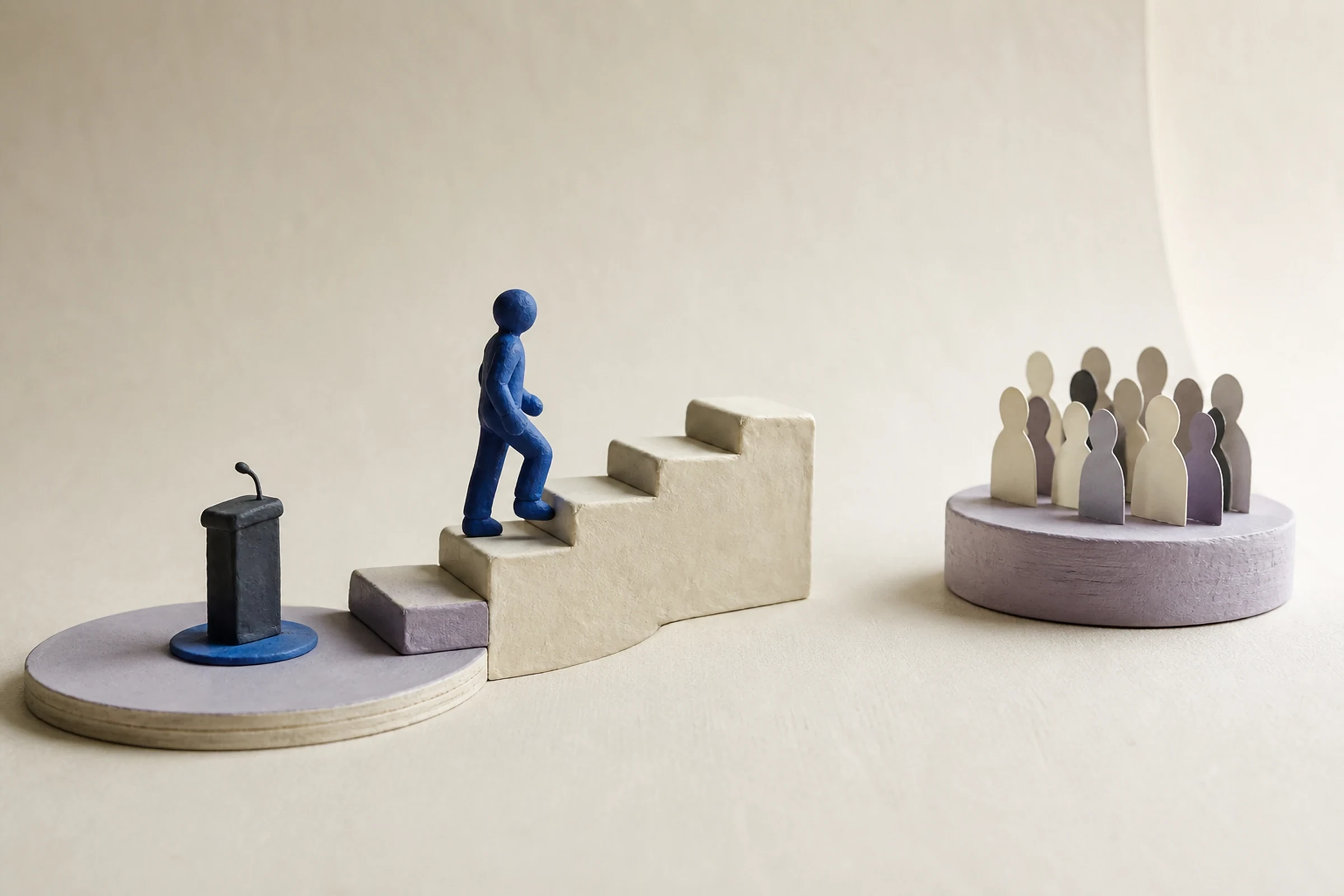 A clay speaker climbs a five-step staircase from a small VR practice stage toward a separate live audience platform, with a visible gap before the final step.
