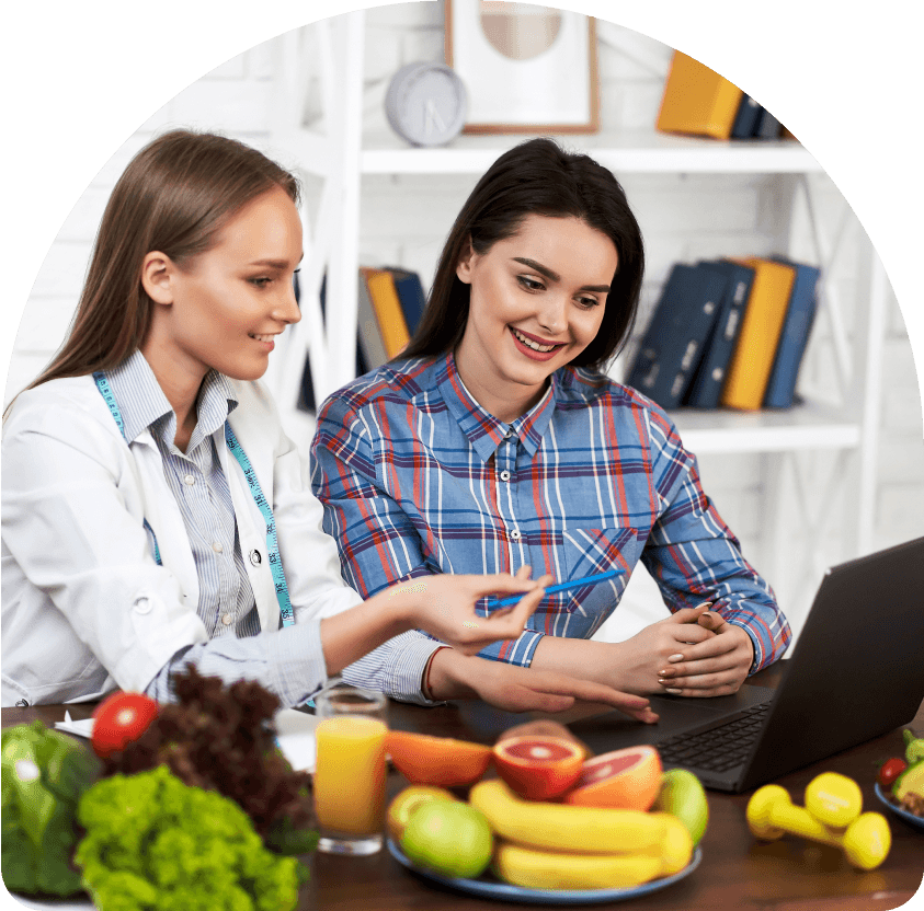 A woman with curly hair sits cross-legged indoors, smiling with her eyes closed, embodying a sense of wellness and relaxation. In the background, another person is also seated. Large windows let in natural light, creating a warm and peaceful atmosphere perfect for mindful introspection and balance.