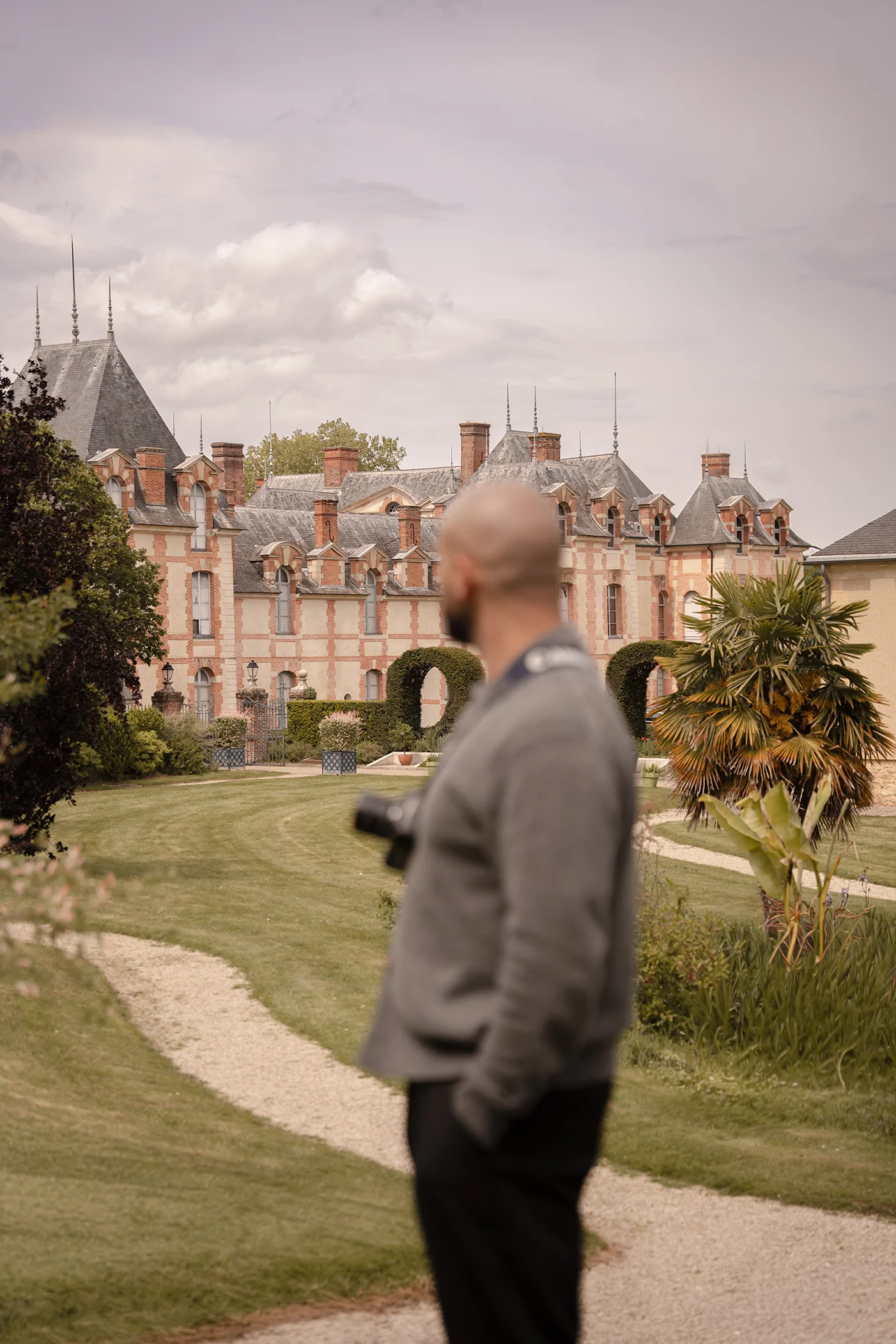 Photographer Quentin Moya seen from behind, camera in hand, looking toward a brick château and its gardens