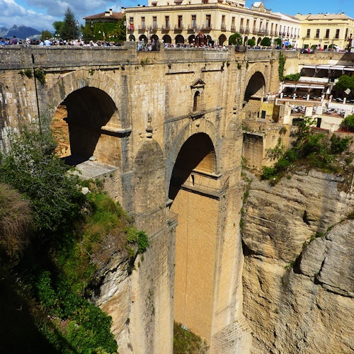 Ancient stone bridge with three large arches spanning a deep gorge, with buildings and people visible in the background.