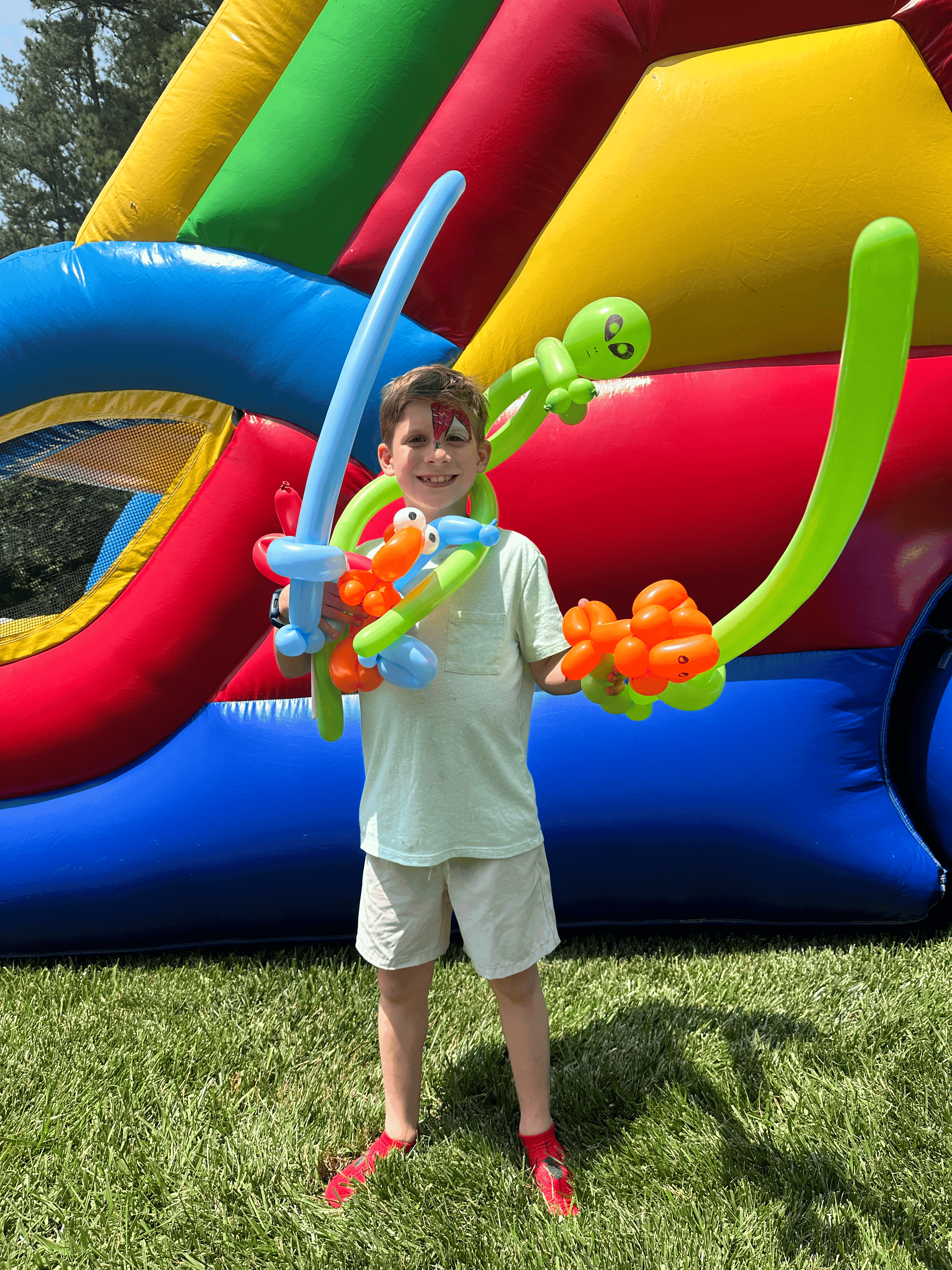 A smiling boy stands in front of a colorful inflatable bounce house, holding an array of balloon animals, including a fish, alien, and sword. He has a small face painting of a red mask, complementing the festive outdoor setting.