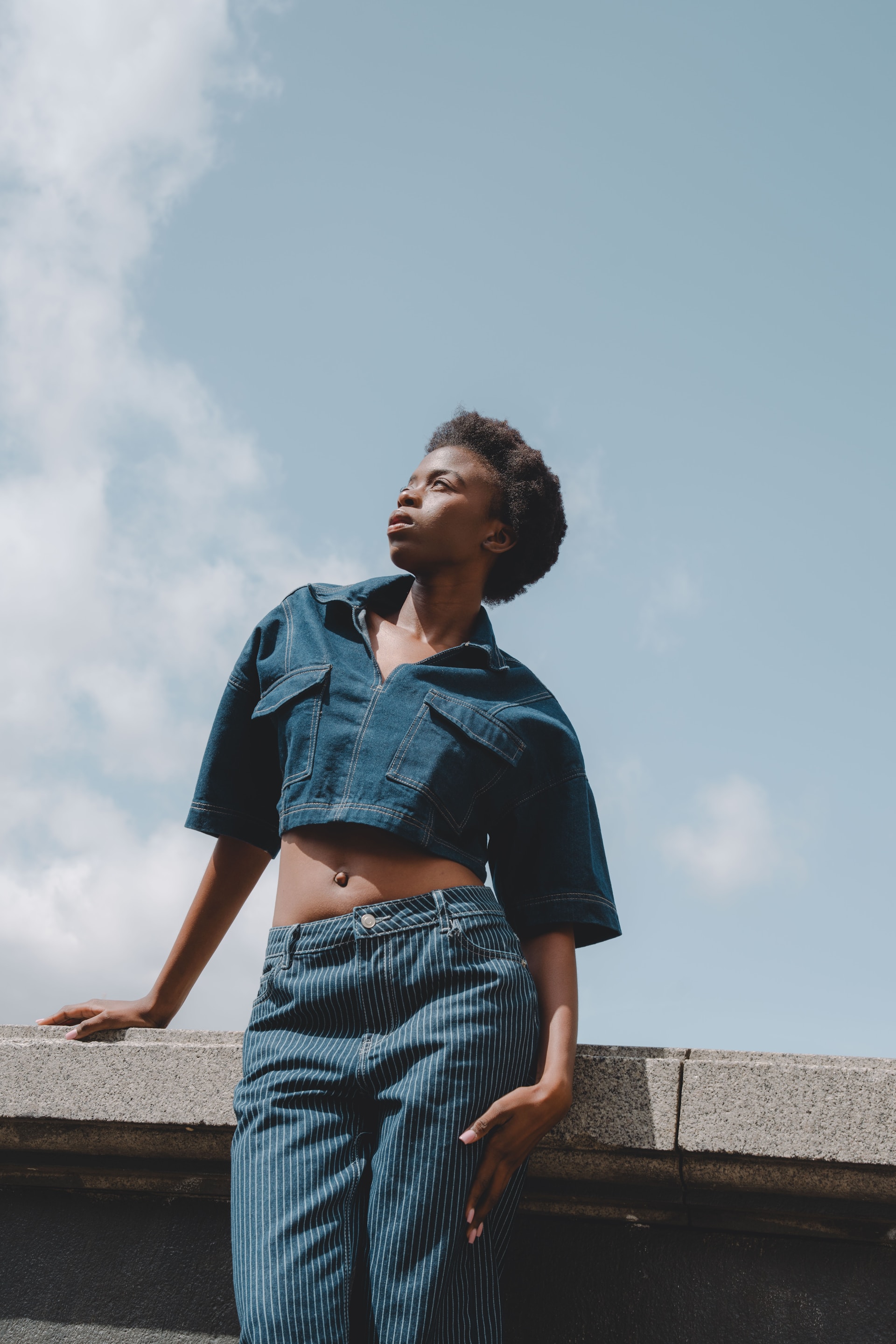 A low-angle shot of the woman in denim looking upward and away from the camera. The bright blue sky and soft white clouds fill the background above the stone ledge she is leaning on.