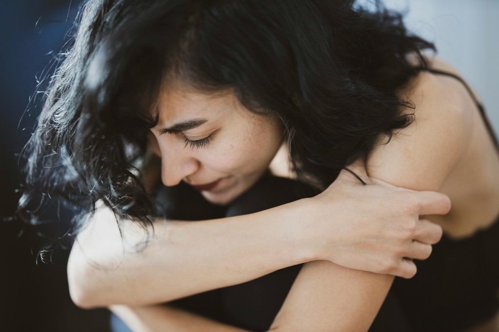 A young woman with dark hair hugs her knees and closes her eyes, portraying a moment of deep emotional distress and introspection.