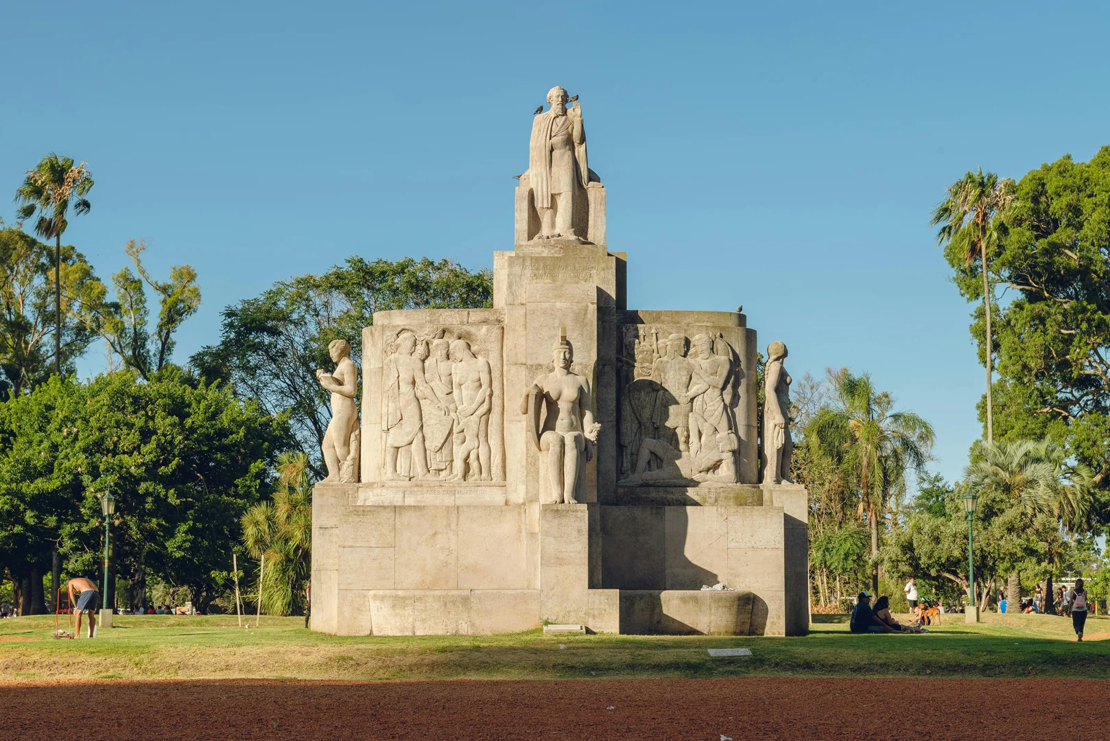 Monumento de piedra en un parque verde urbano, rodeado de árboles bajo un cielo azul despejado.