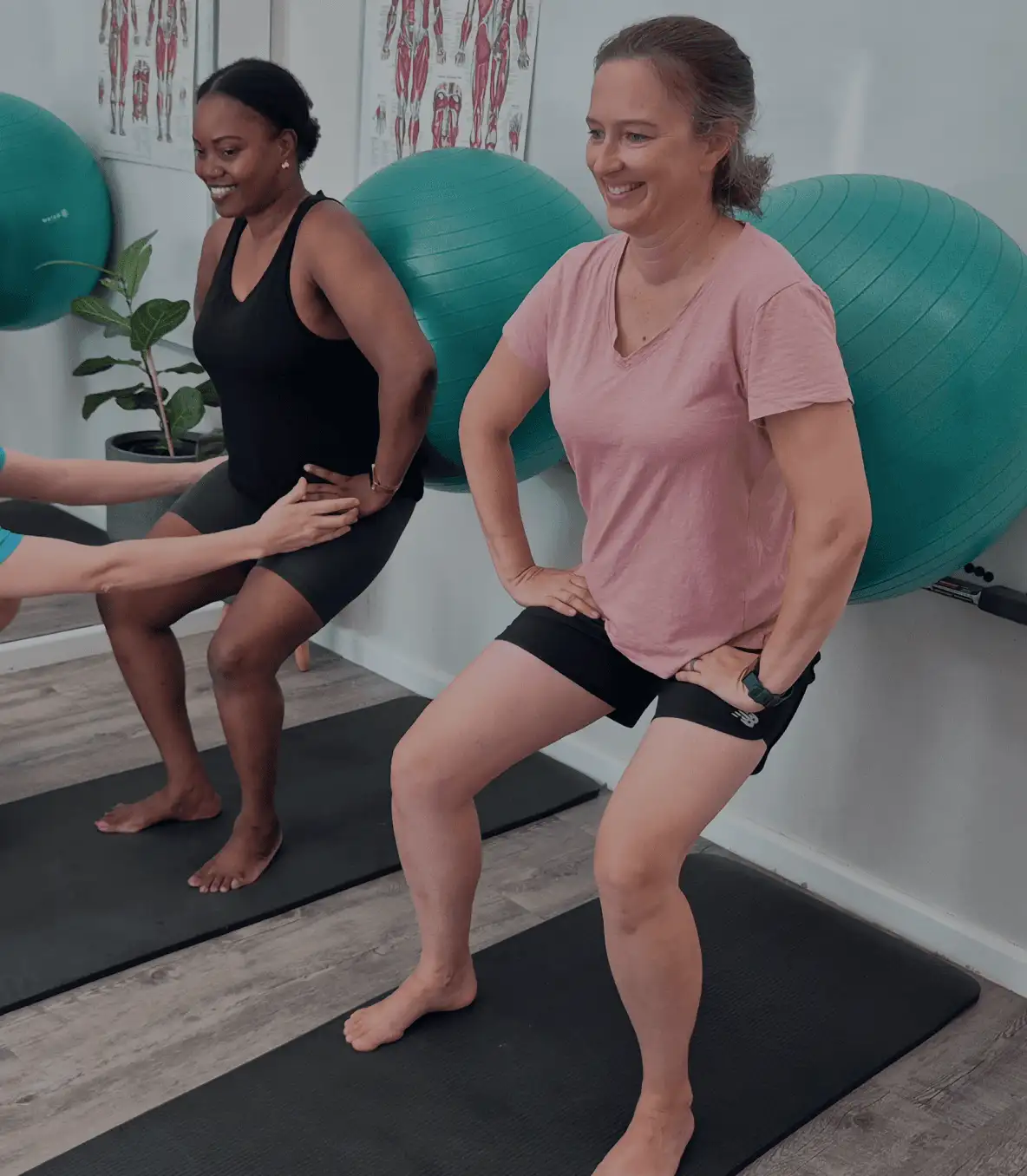 Two women in an exercise class performing ball squats in a seated position against the wall in a gym.