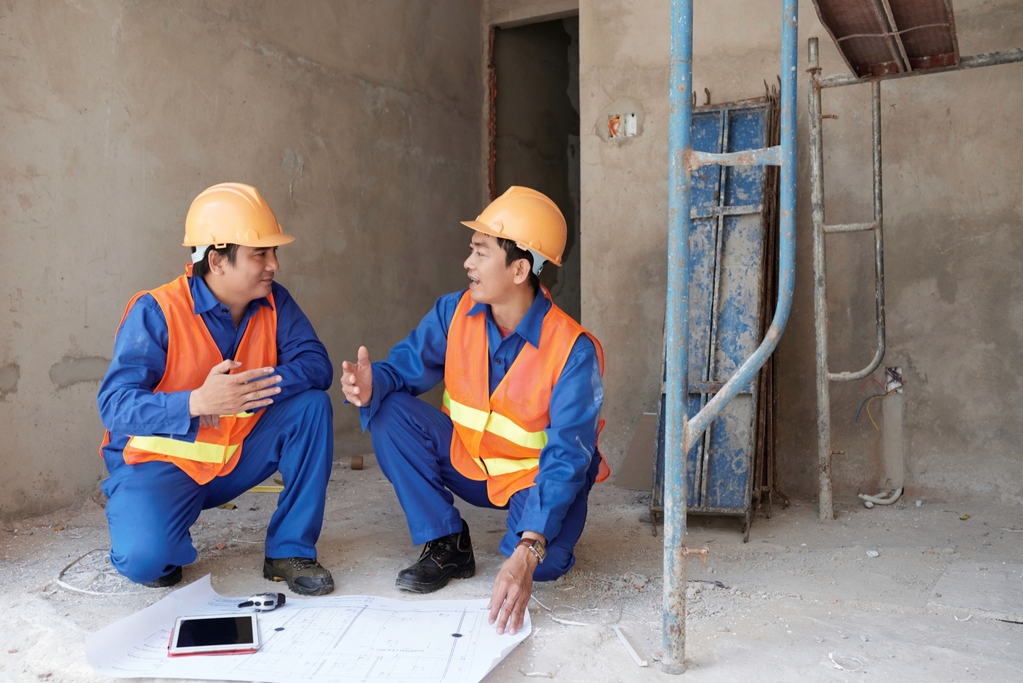 兩名穿著安全帽及橙色反光衣的建築工人在工地核對圖則，配合建築工程保險及中小企裝修保險。