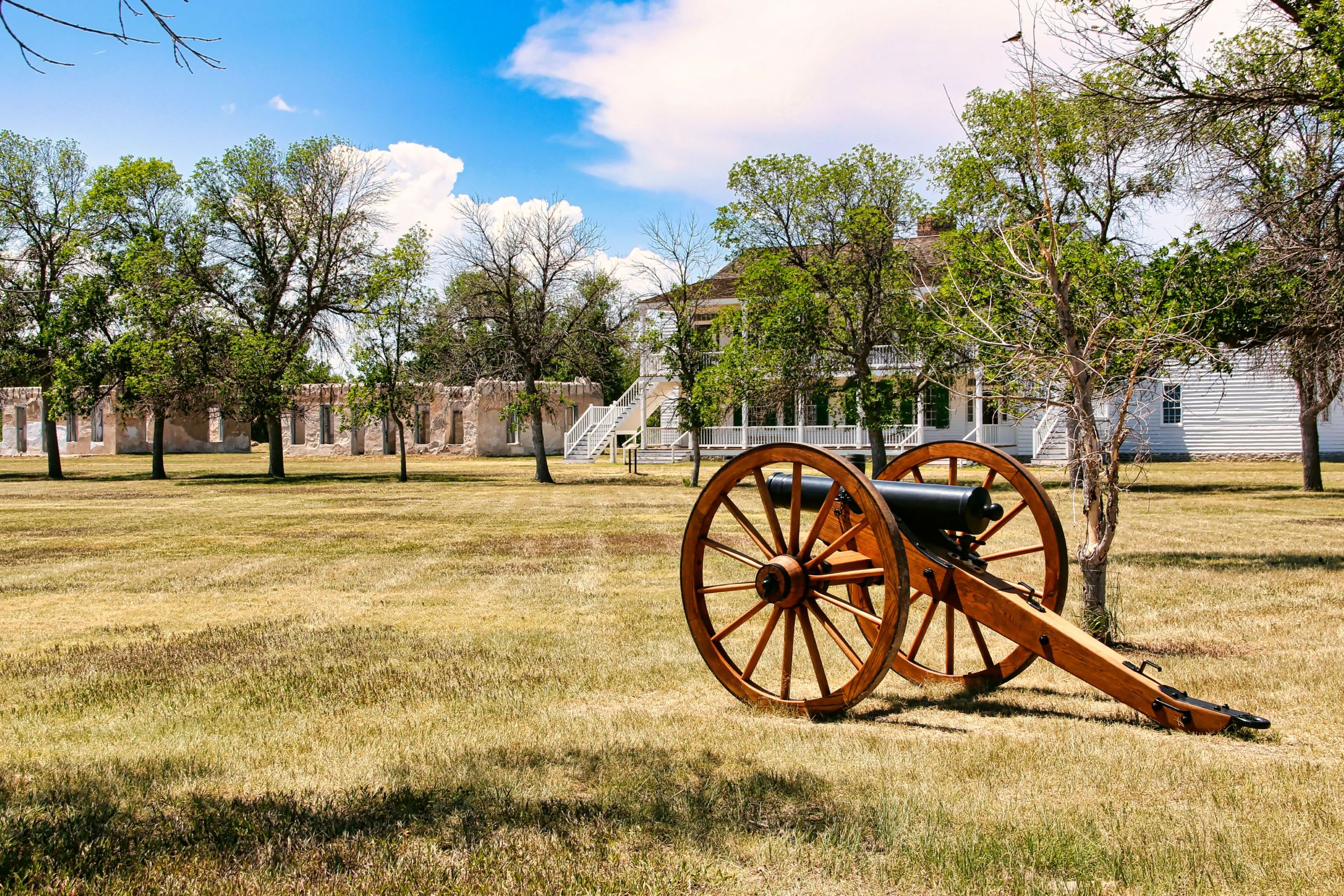 A wooden cannon sits on a grassy field in front of historic barracks buildings at Fort Laramie National Historic Site.