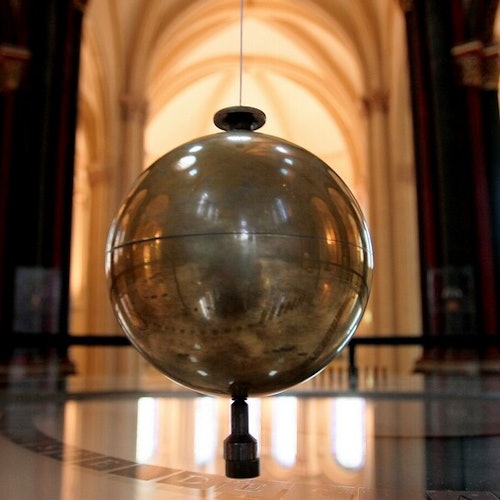 Un gran péndulo brillante colgando dentro de un majestuoso edificio con techos arqueados y columnas ornamentadas al fondo.