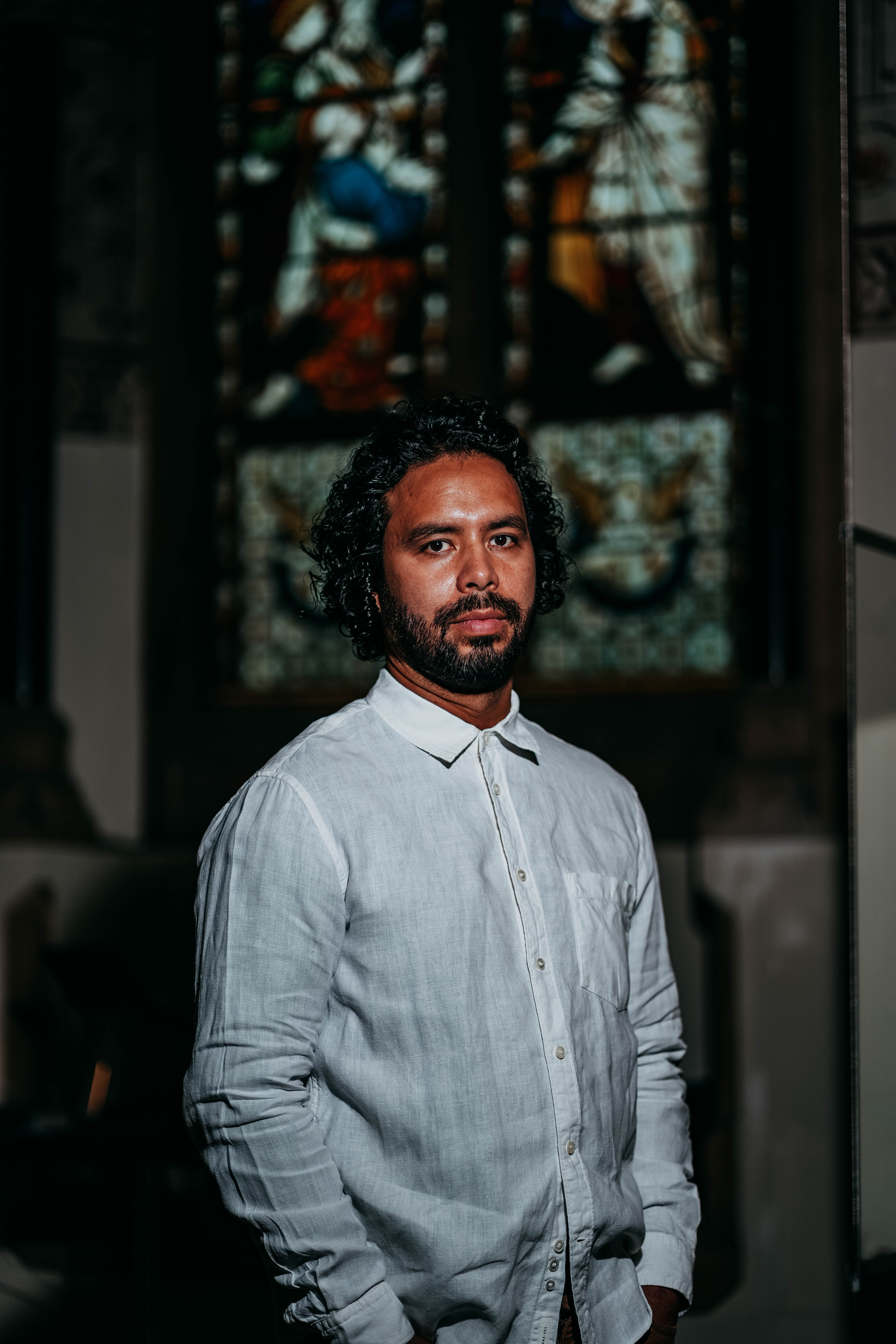 A close-up, side-profile portrait of a man with dark curly hair and a beard looking over his shoulder at the camera. The dramatic lighting illuminates one side of his face against a dark background.
