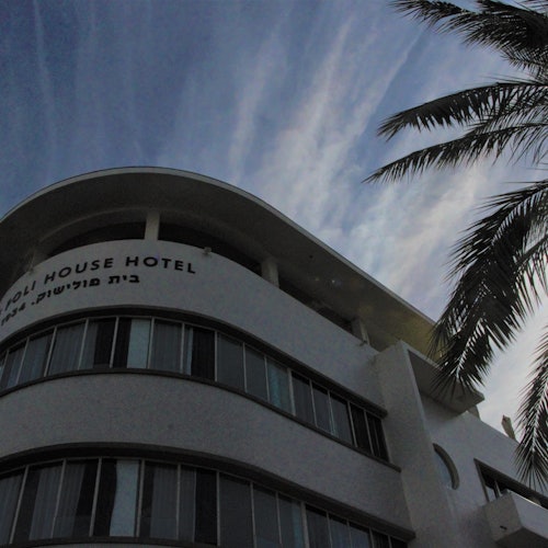 Curved facade of a building labeled "Roll House Hotel" with palm tree leaves in the foreground against a partly cloudy sky.