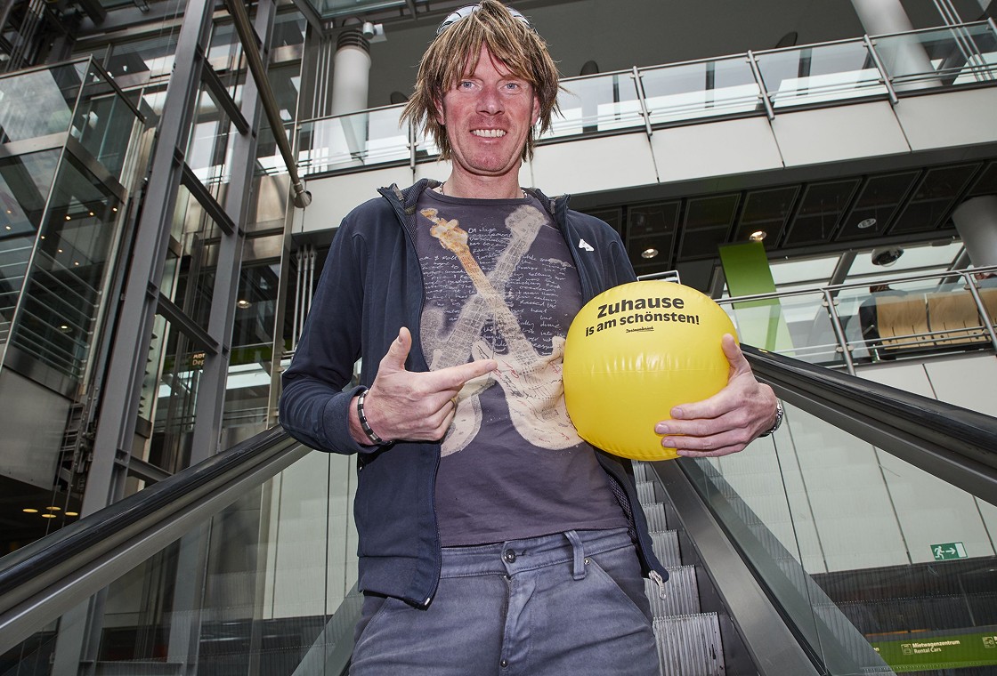 Mickie Krause steht mit gelbem Ball auf einer Rolltreppe im Flughafen Dortmund