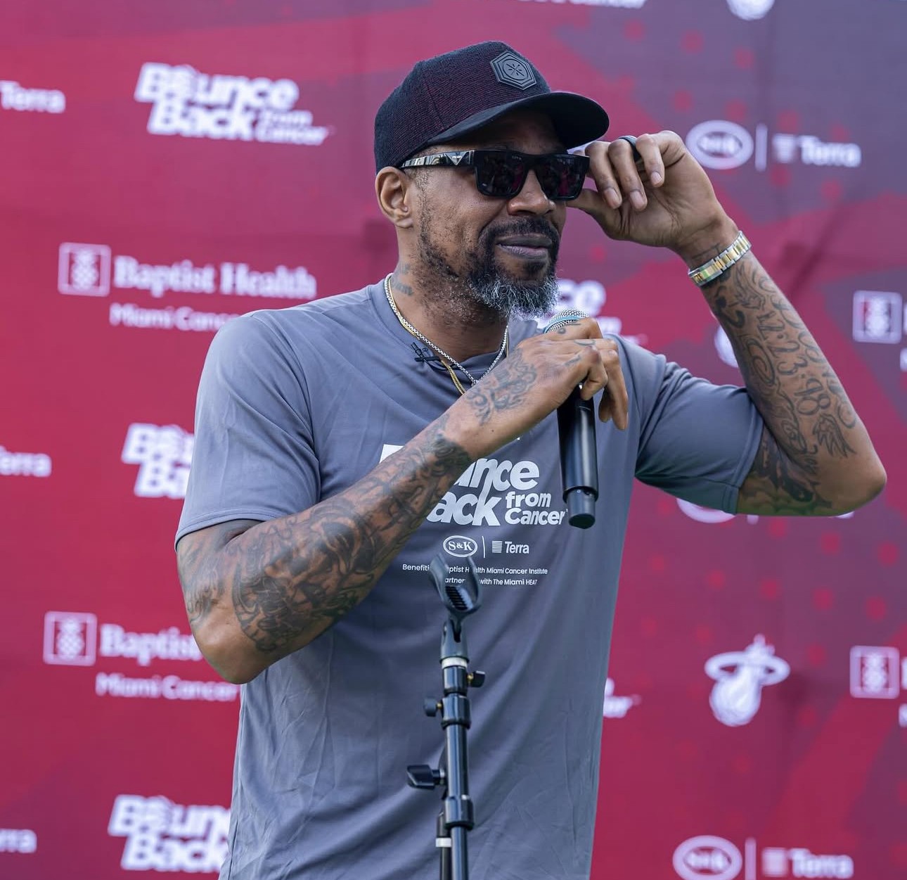 Udonis Haslem wearing sunglasses and a cap speaks into a microphone. He is wearing a gray Bounce Back from Cancer shirt, with a red backdrop behind him featuring Baptist Health and Miami Heat logos.
