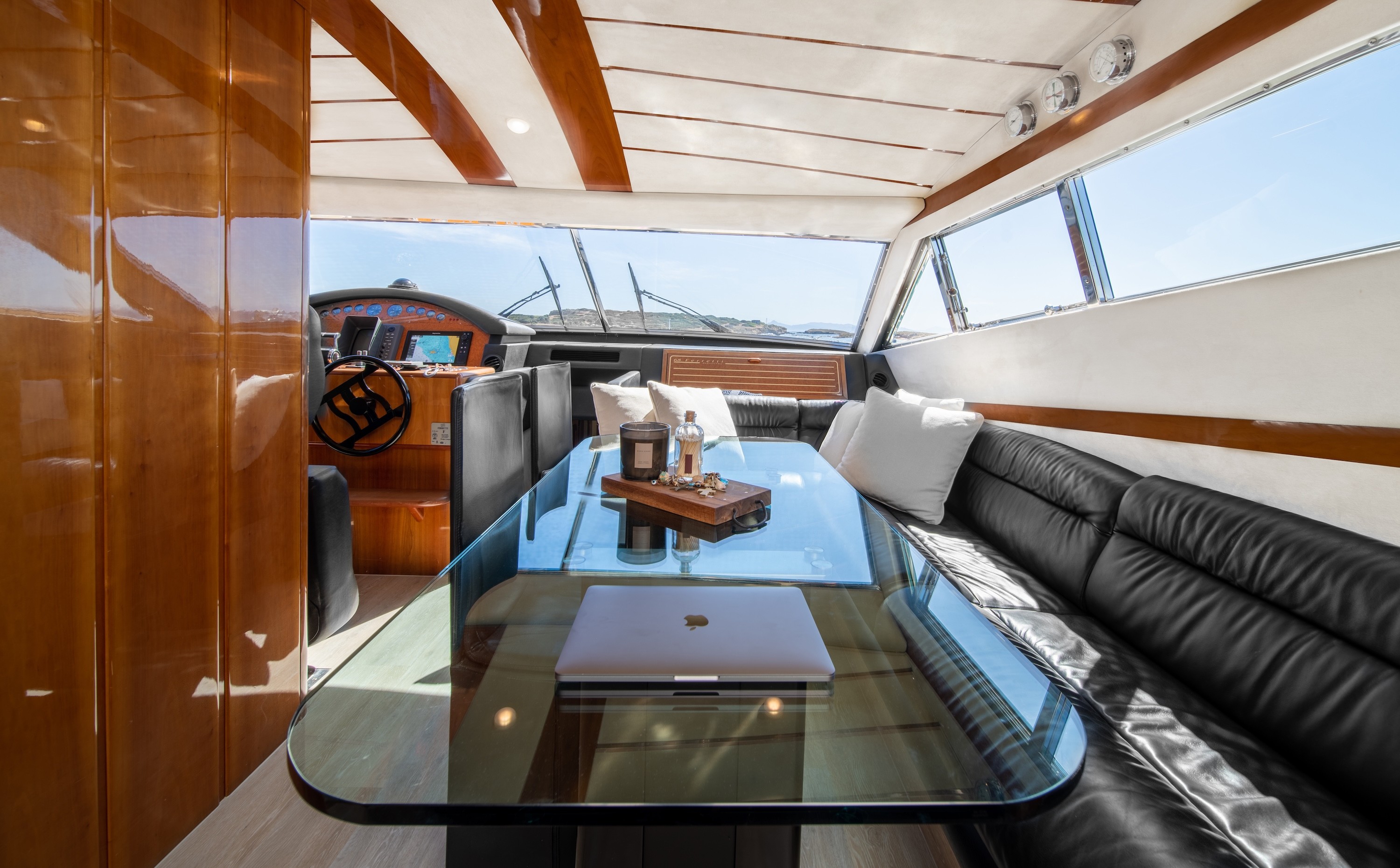 Luxury yacht salon interior featuring white leather seating, polished wood cabinetry, glass coffee table with plant, and flat-screen TV with marina views through large windows.