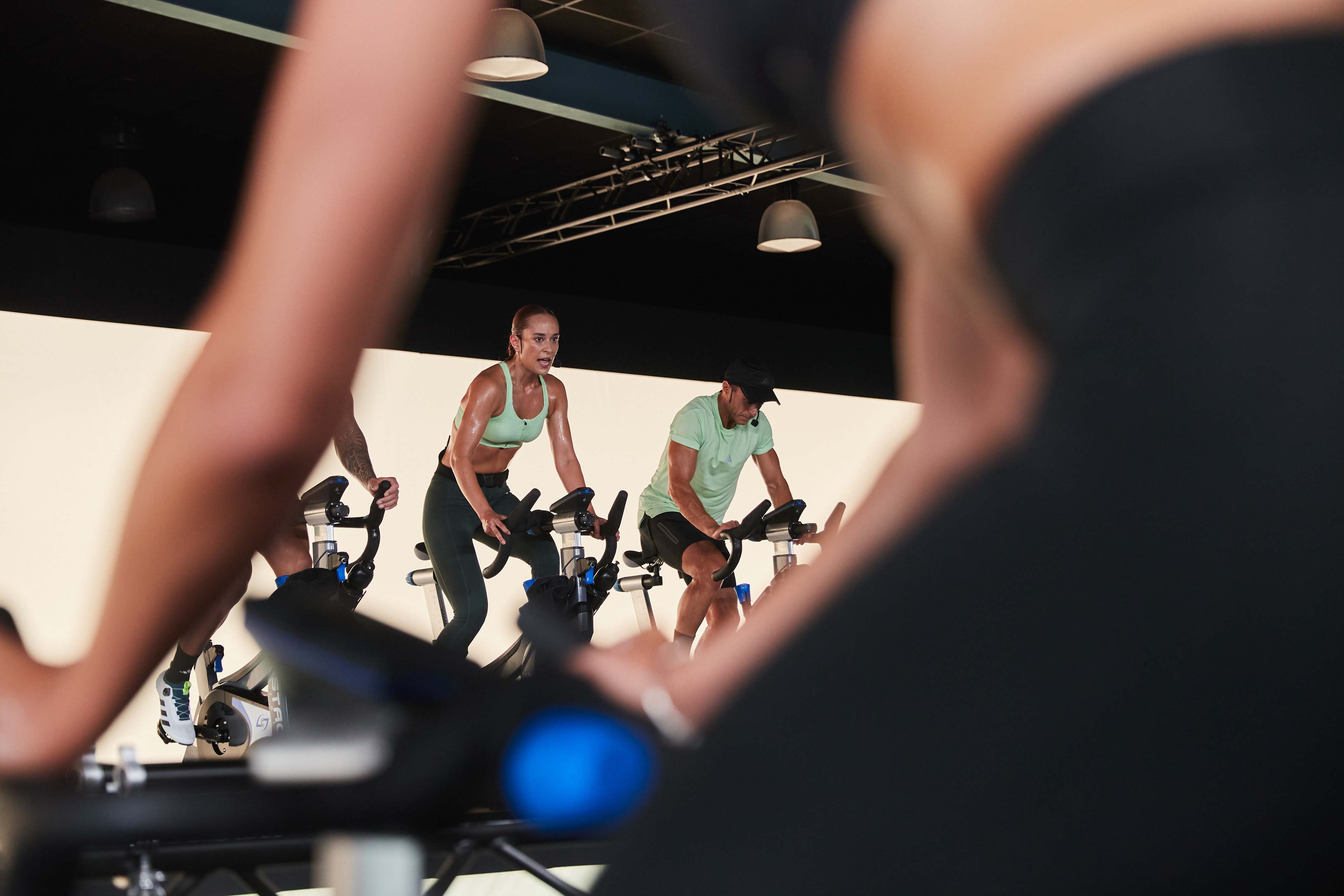 Teilnehmer und Trainer in einer energiegeladenen Group Cycling-Klasse auf Indoor-Bikes. Eine Trainerin im Vordergrund ist fokussiert und schweißgebadet, während andere Teilnehmer im Hintergrund ebenfalls intensiv trainieren. Die Szene ist in einem modernen Fitnessstudio mit schlichtem Hintergrund aufgenommen.