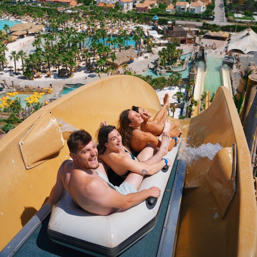 Three people on a water slide raft, smiling and enjoying the ride. The background shows a water park with pools and tropical scenery.