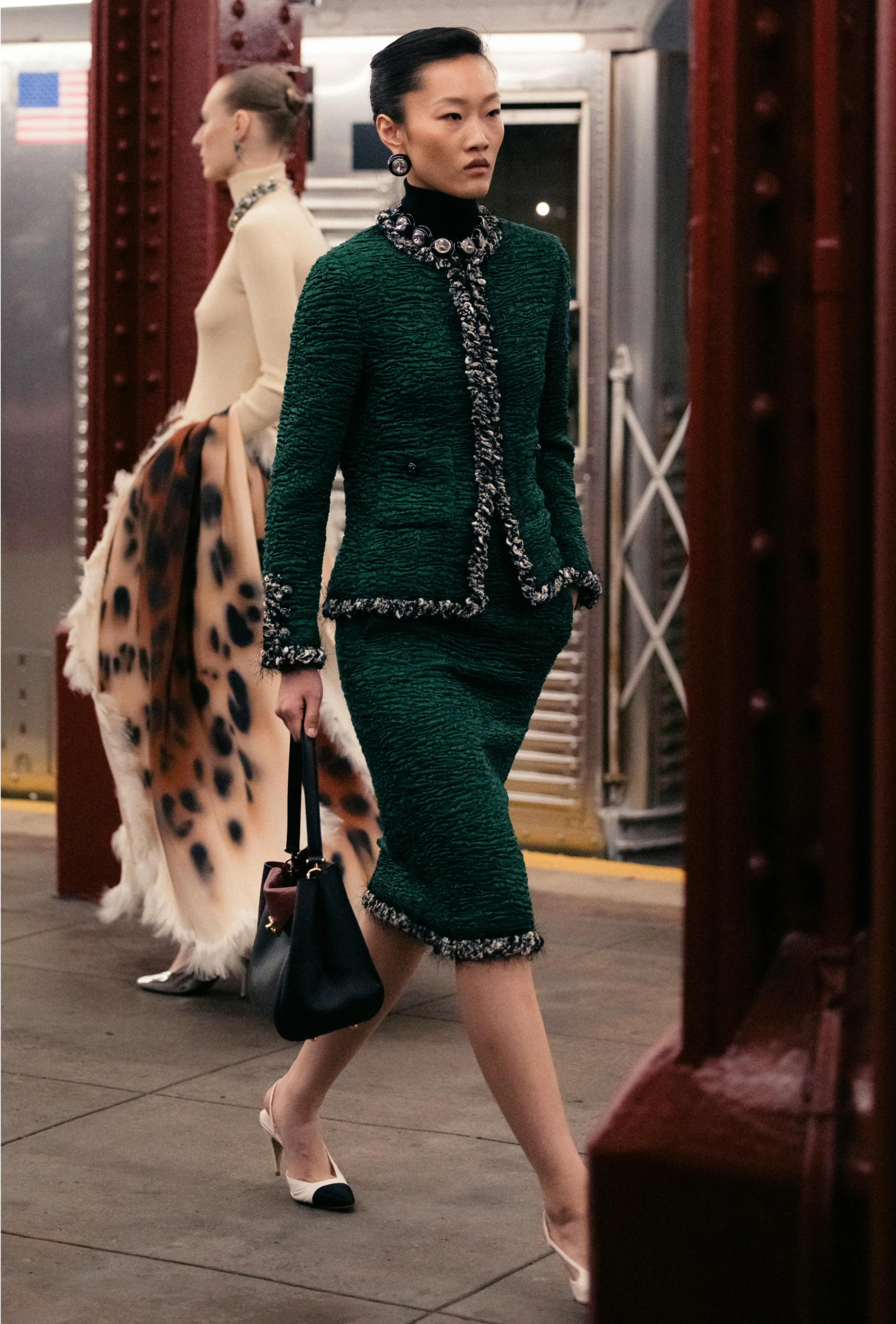 A model in a dark green tweed skirt suit with black trim walks across a subway platform carrying a handbag at the Chanel Métiers d’Art 2026 show.