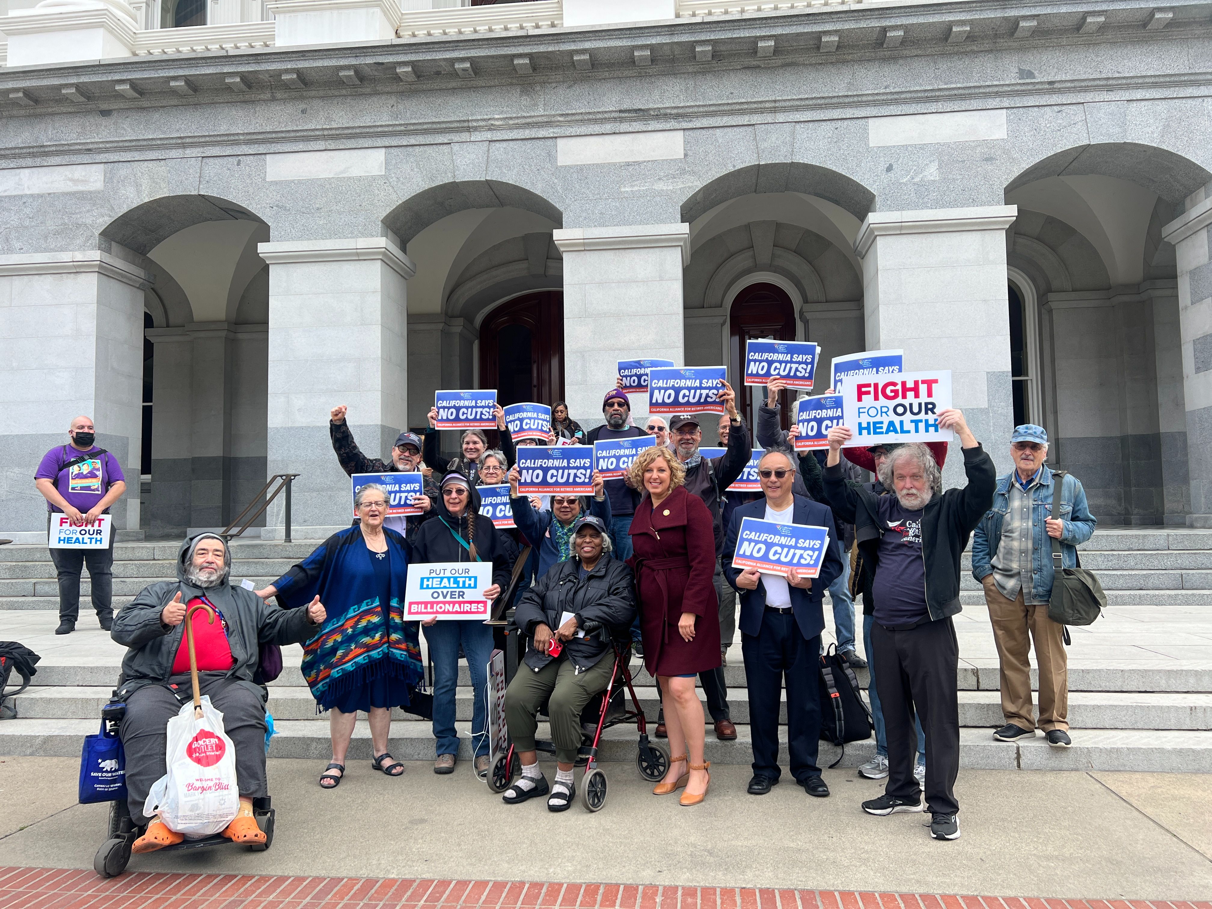 A group of advocates, including individuals using mobility aids, stand on the steps of a government building holding signs that read “NO CUTS” and “FIGHT FOR OUR HEALTH,” showing support for healthcare protections.