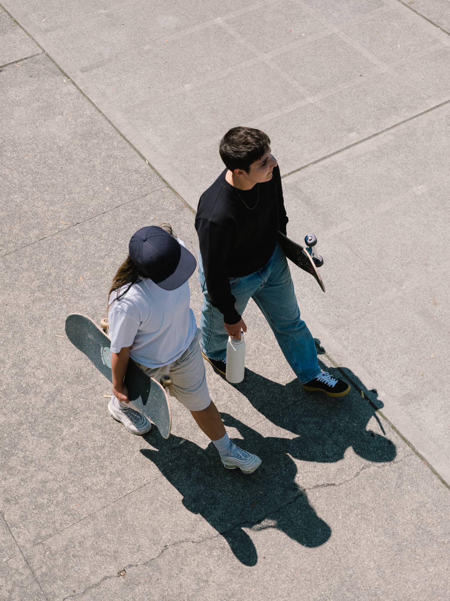 A person in a black outfit and red sneakers stands confidently on stacked wooden planks with a skateboard. Background features lush greenery.