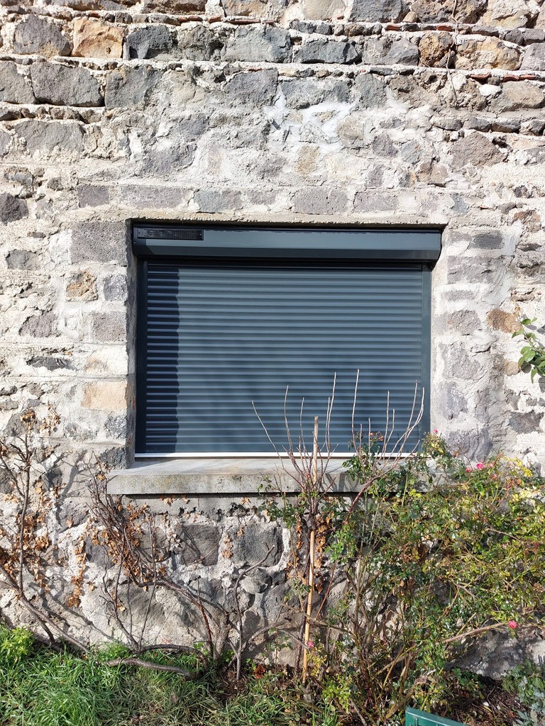 Mur en pierre ancienne avec volet roulant SOMFY IO gris fermé.