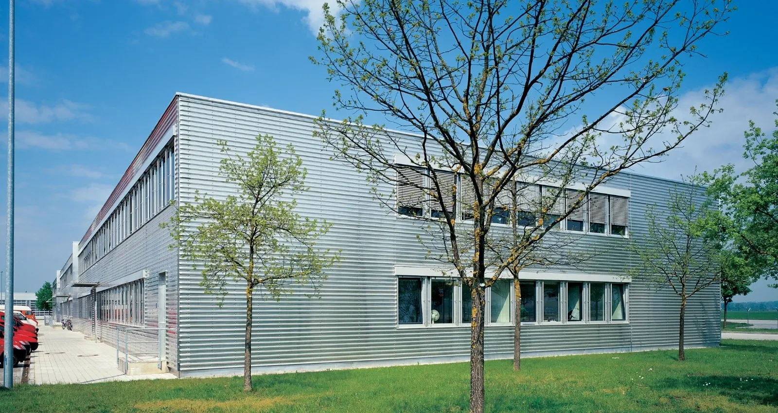 Moderne Fabrik mit geriffelter Metallfassade und großzügigen Fensterfronten, umgeben von Rasenflächen und jungen Bäumen.