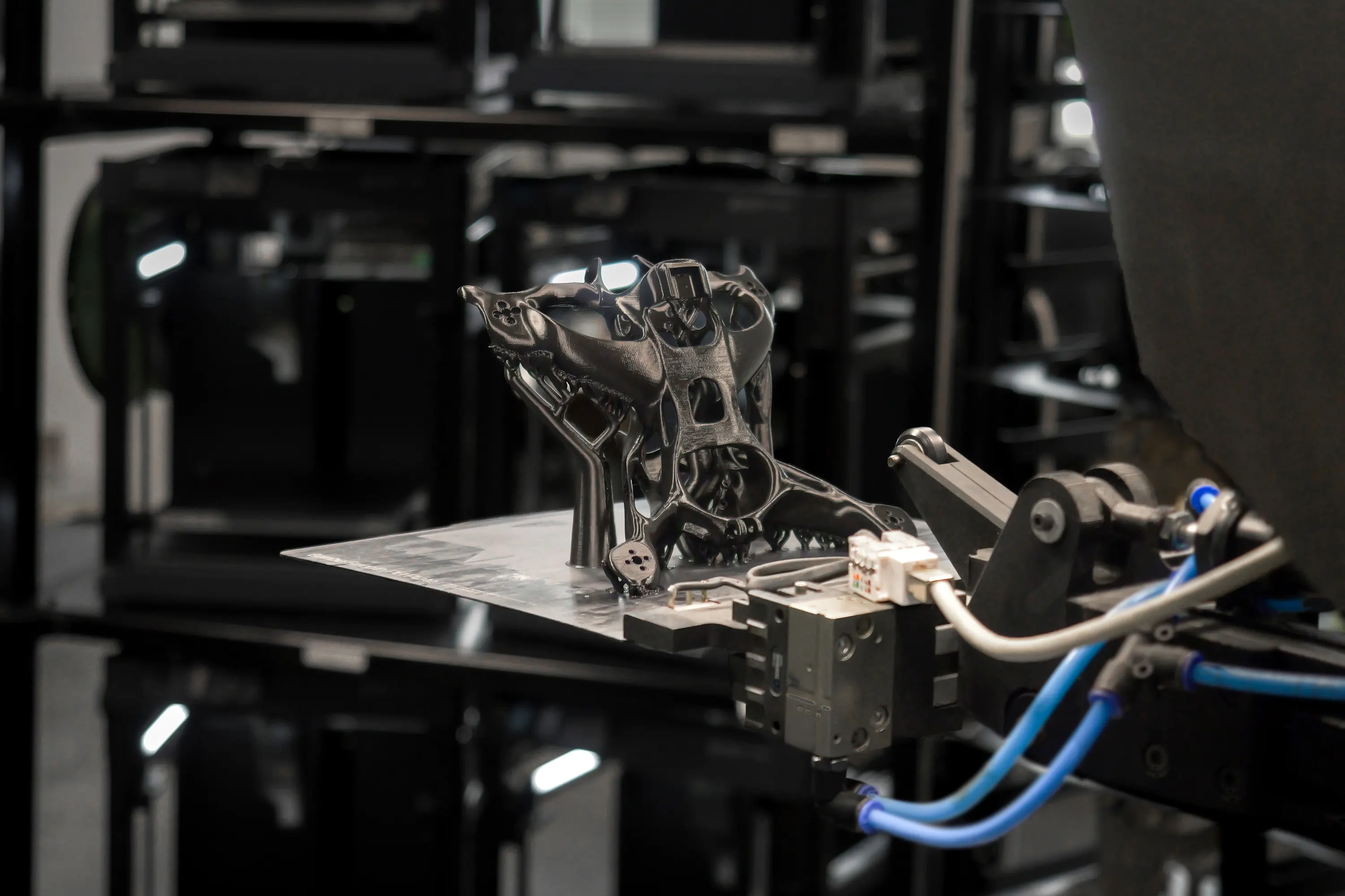 A robotic arm intricately handles a black 3D-printed drone airframe in a manufacturing facility, showcasing advanced robotic automation and innovative plate handling within a 3D print farm, highlighting its potential use in drone manufacturing.