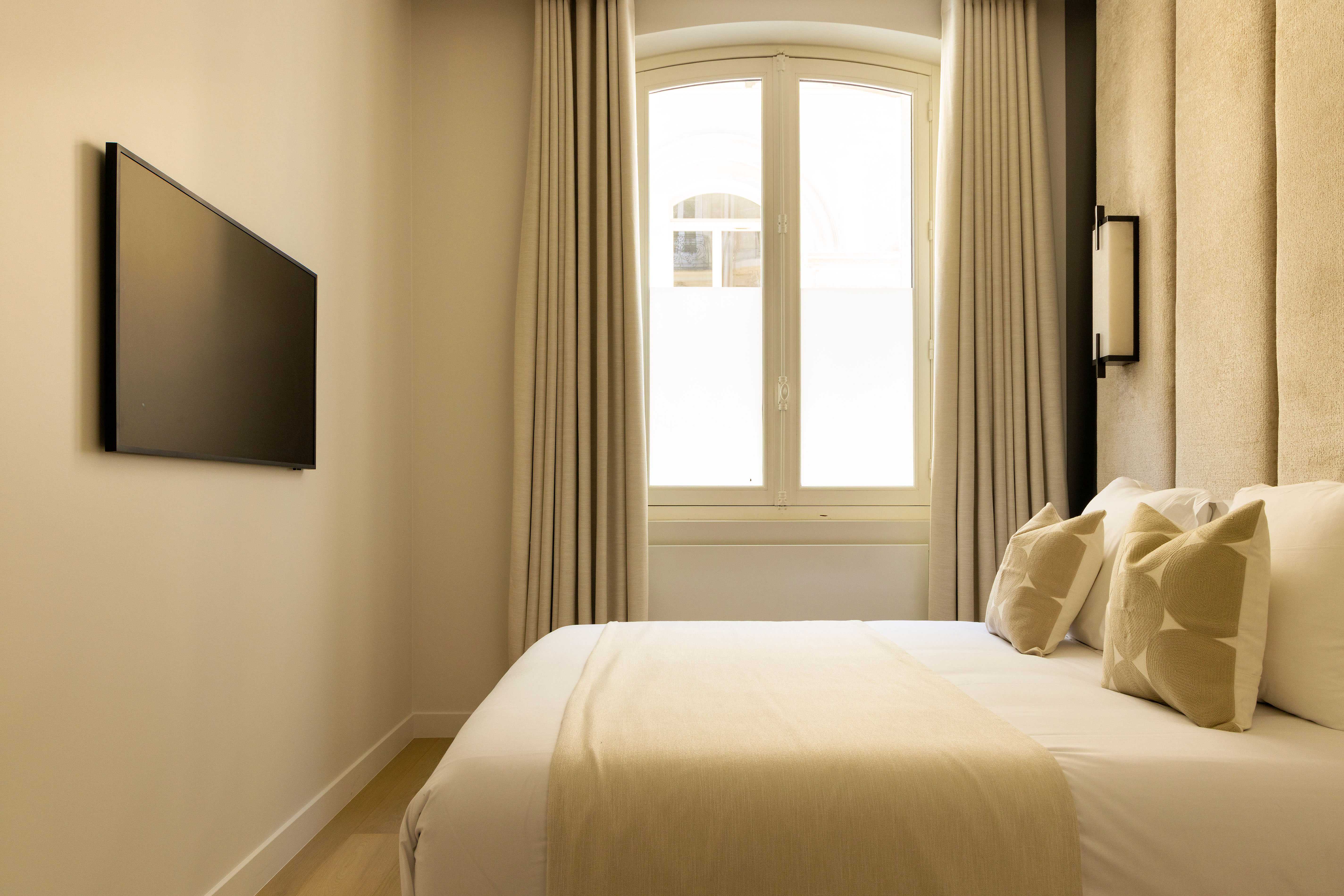 Chambre avec fenêtre cintrée, lit blanc, télévision murale et tête de lit velours beige