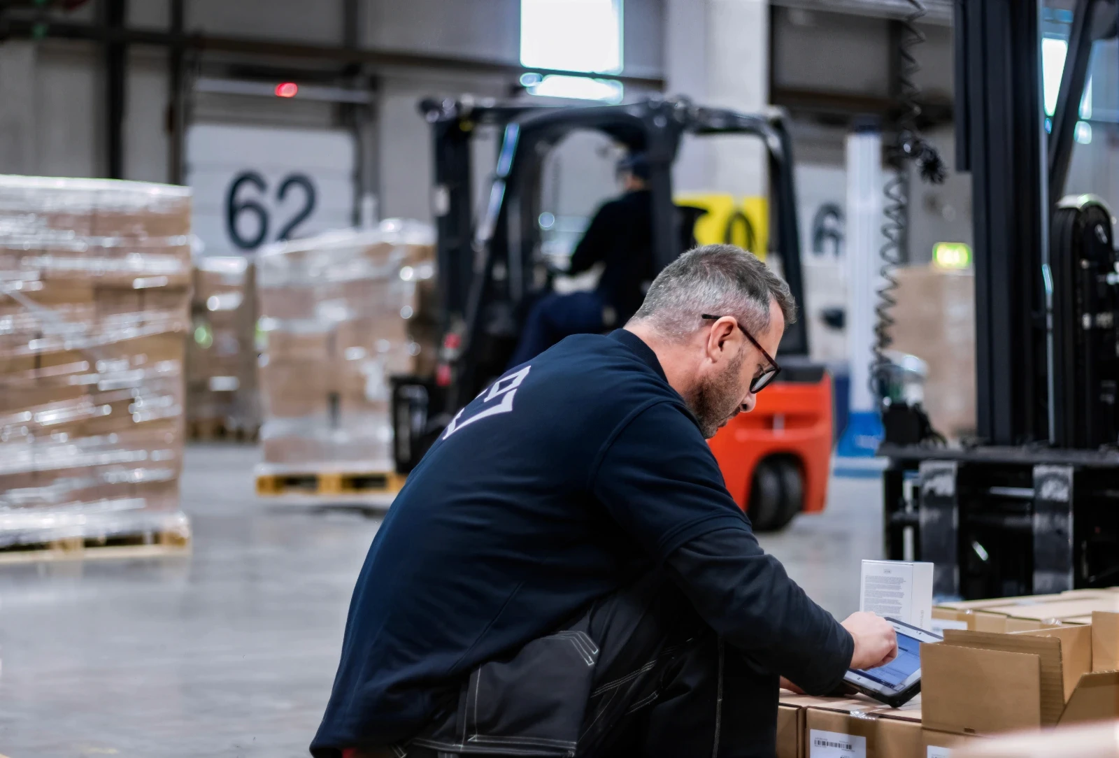 Man i lager klädd i arbetskläder använder surfplatta medan gaffeltrucken jobbar.