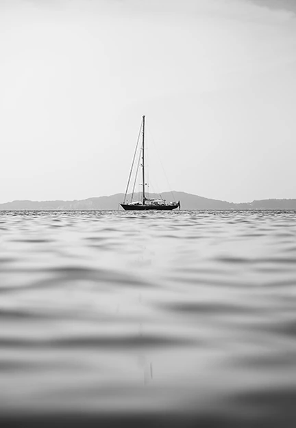 Czarno-białe zdjęcie samotnego jachtu żaglowego na spokojnym morzu z rozmytą taflą wody na pierwszym planie