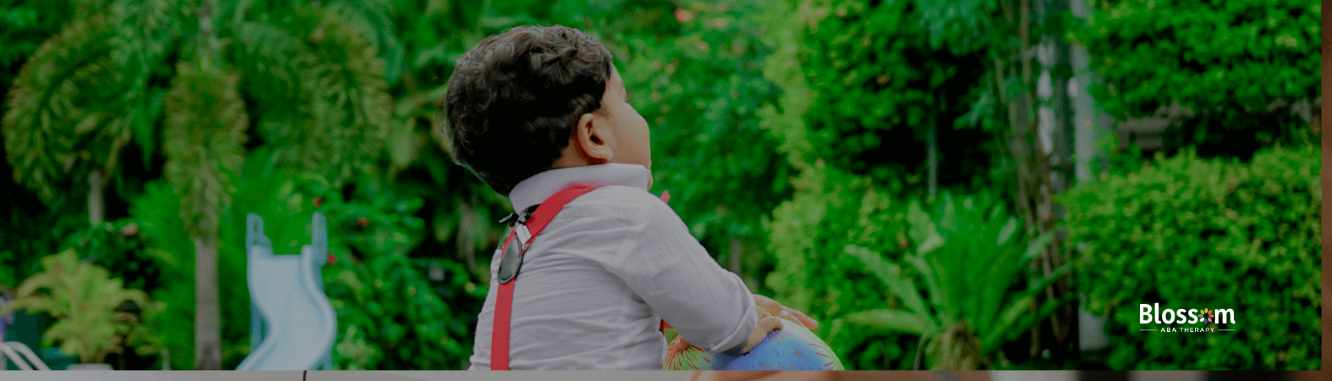 Young child sitting near a pool holding a colorful ball, surrounded by bright green plants and a relaxed outdoor setting.