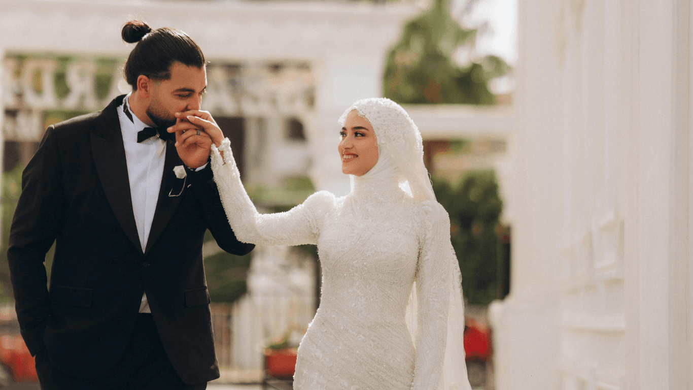 A couple in formal attire walking hand-in-hand, smiling, in an elegant outdoor setting. The woman wears a hijab.