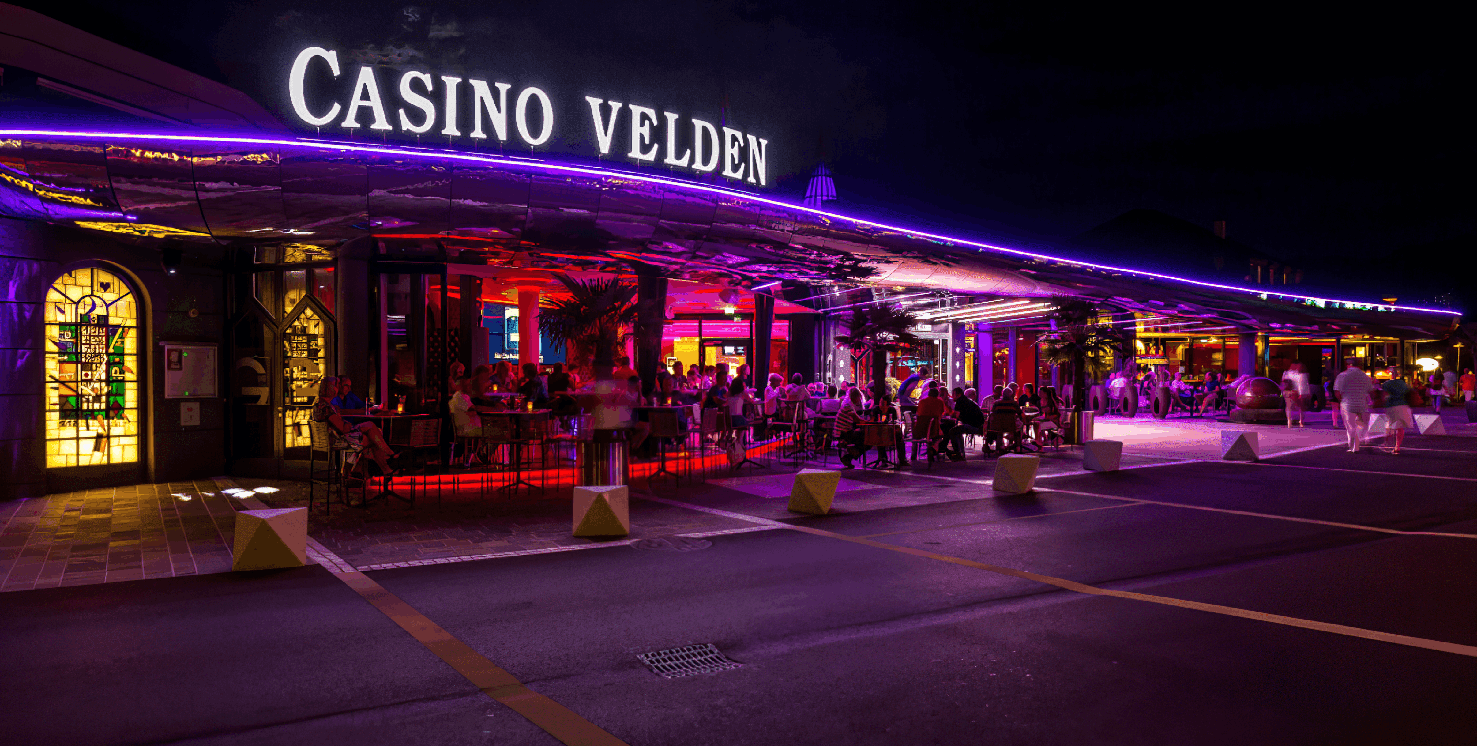 MW Interior  - Casino Velden am Wörthersee bei Nacht mit violetter LED-Beleuchtung und belebter Außenterrasse" Beschreibung: Das Casino Velden am Wörthersee – ein anspruchsvoller Referenzstandort, für den hochwertige Einrichtungslösungen im Hospitality-Bereich realisiert wurden.