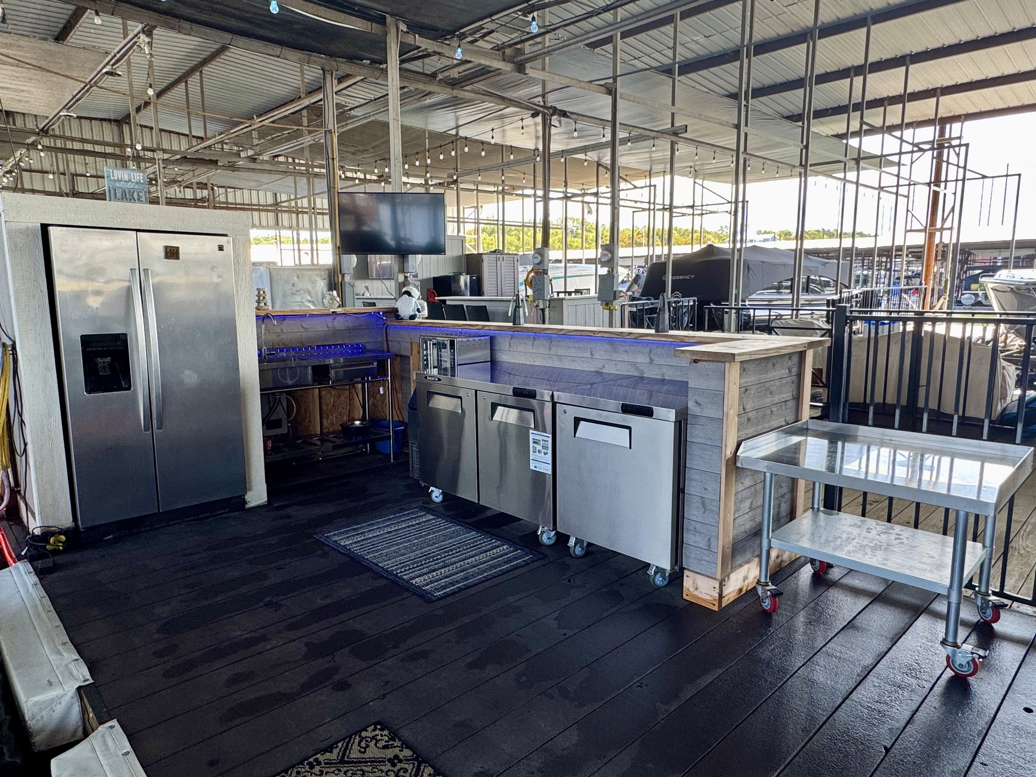 Spacious outdoor kitchen area with a large stainless steel refrigerator, double-door coolers, a set of shelves, and a mounted television under a metal-roofed open structure.